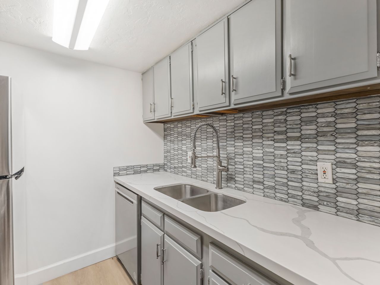 Kitchen featuring light gray cabinetry with brushed nickel hardware, a stainless steel dual basin sink with a gooseneck faucet, white stone-finish countertops with veining, and a mosaic tile backsplash