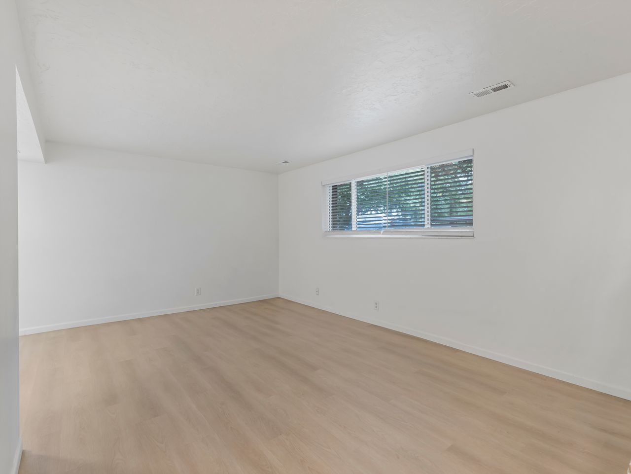 Spacious room featuring wood-finish flooring, white walls, and a large window with horizontal blinds