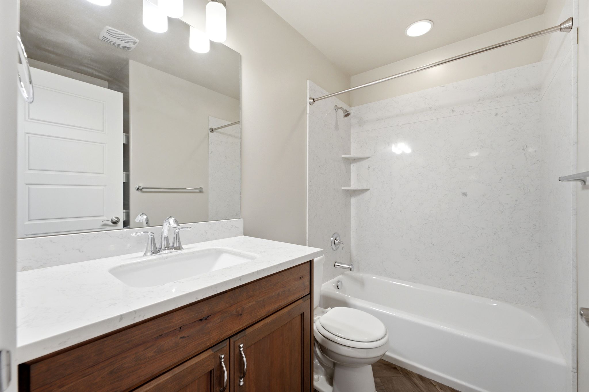 Bathroom featuring a dark wood-finish vanity with a white countertop and integrated sink, a rectangular mirror, a three-light vanity fixture, a white toilet, and a white tub with a white surround