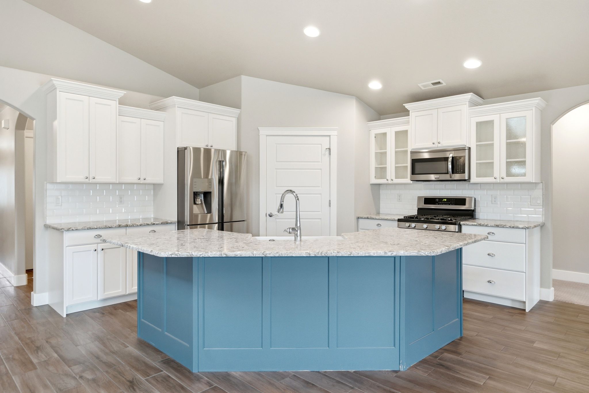 Spacious kitchen featuring a blue central island with a sink, white shaker-style cabinetry, stainless steel appliances, speckled countertops, and wood-finish flooring