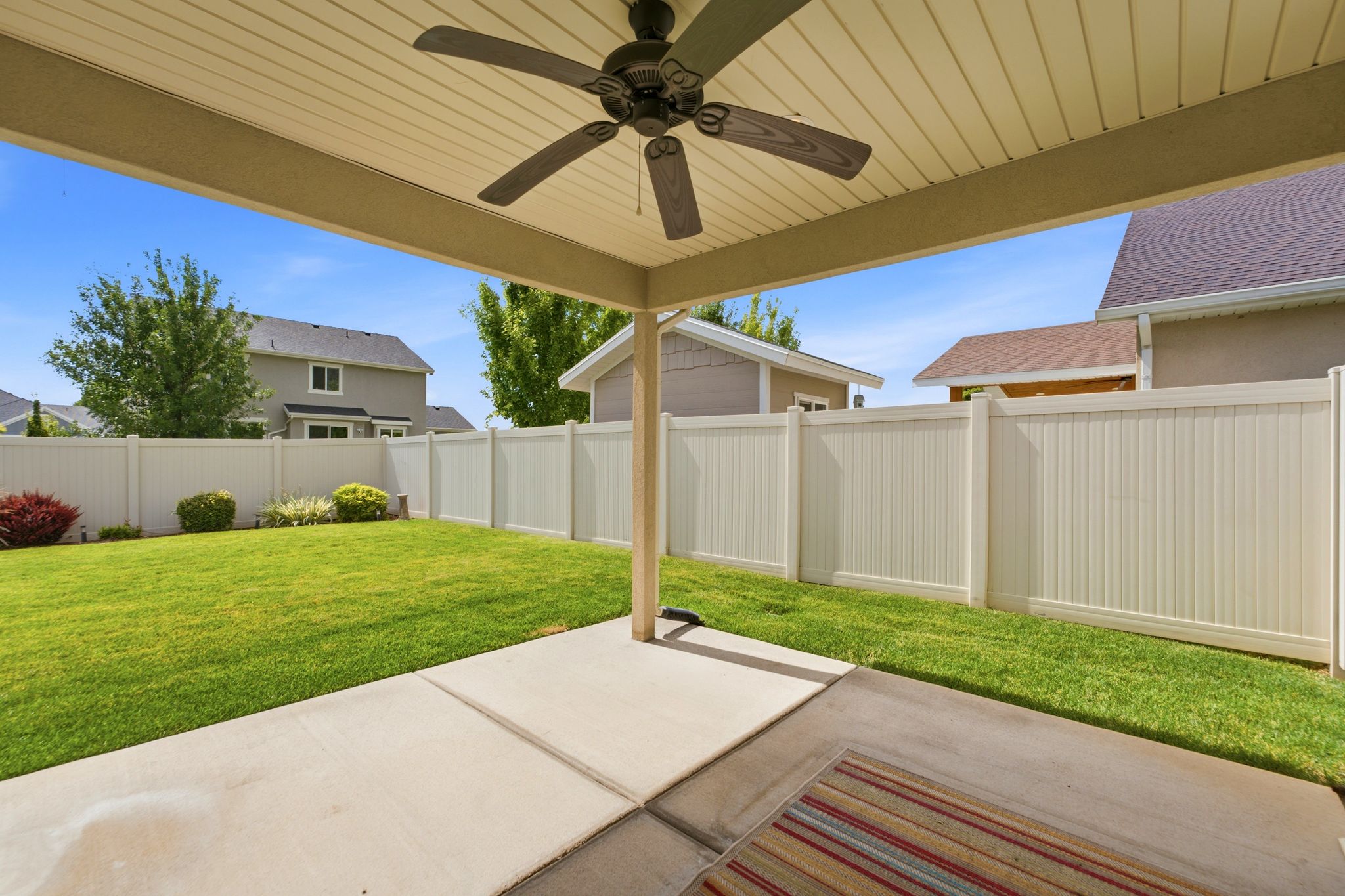 Covered patio area with a ceiling fan, concrete slab, and a fully fenced backyard with a manicured lawn and mature shrubs