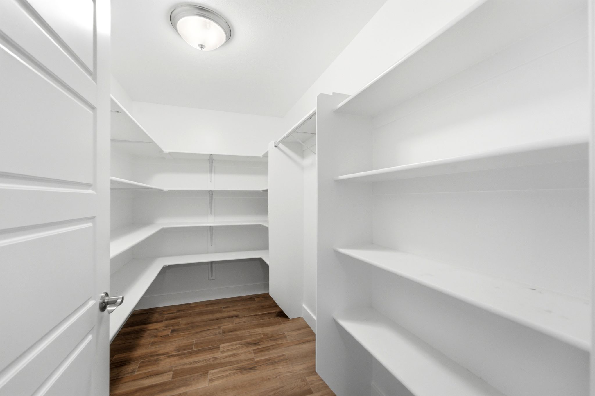 Spacious walk-in pantry featuring extensive white shelving, wood-finish flooring, and a flush-mount ceiling light fixture