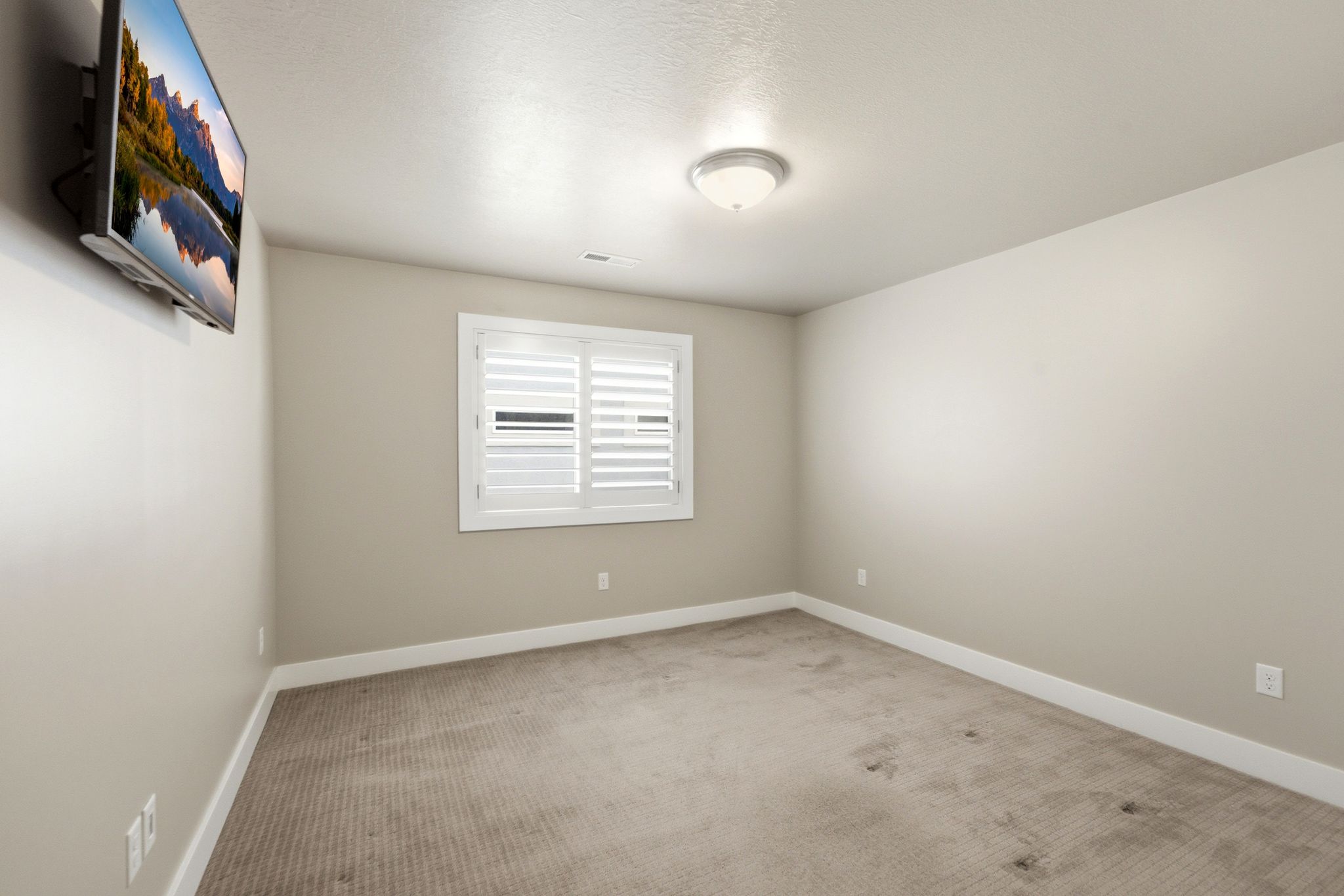 Neutral-toned room featuring a single window with white plantation shutters, an overhead flush-mount light fixture, and textured carpeting
