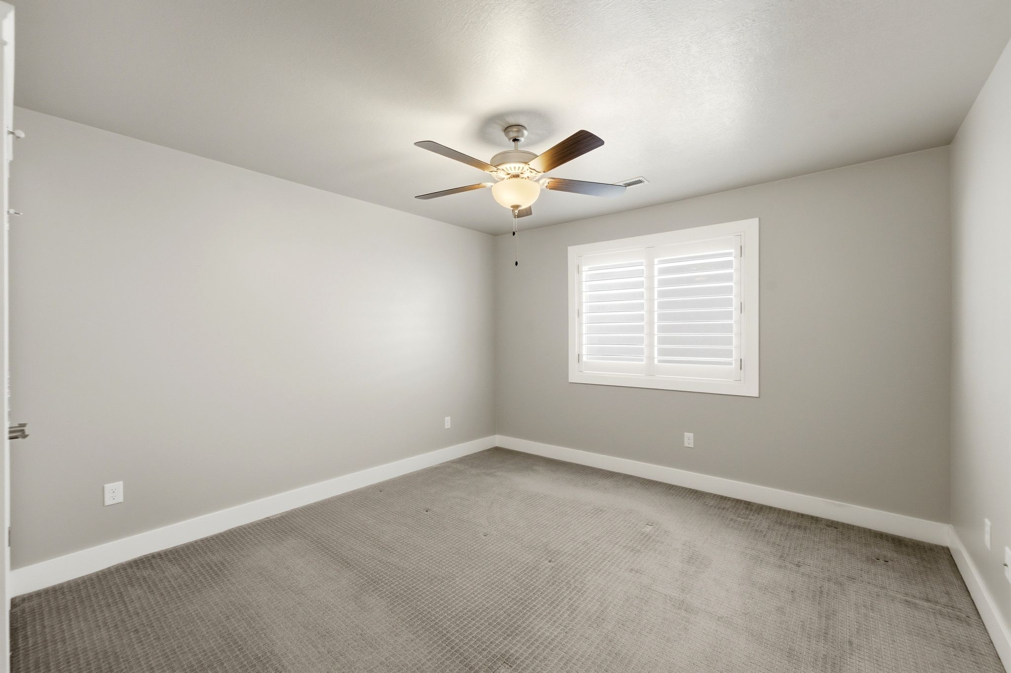 Spacious room featuring neutral-toned walls, plush carpet flooring, a ceiling fan with integrated lighting, and a window with white plantation shutters