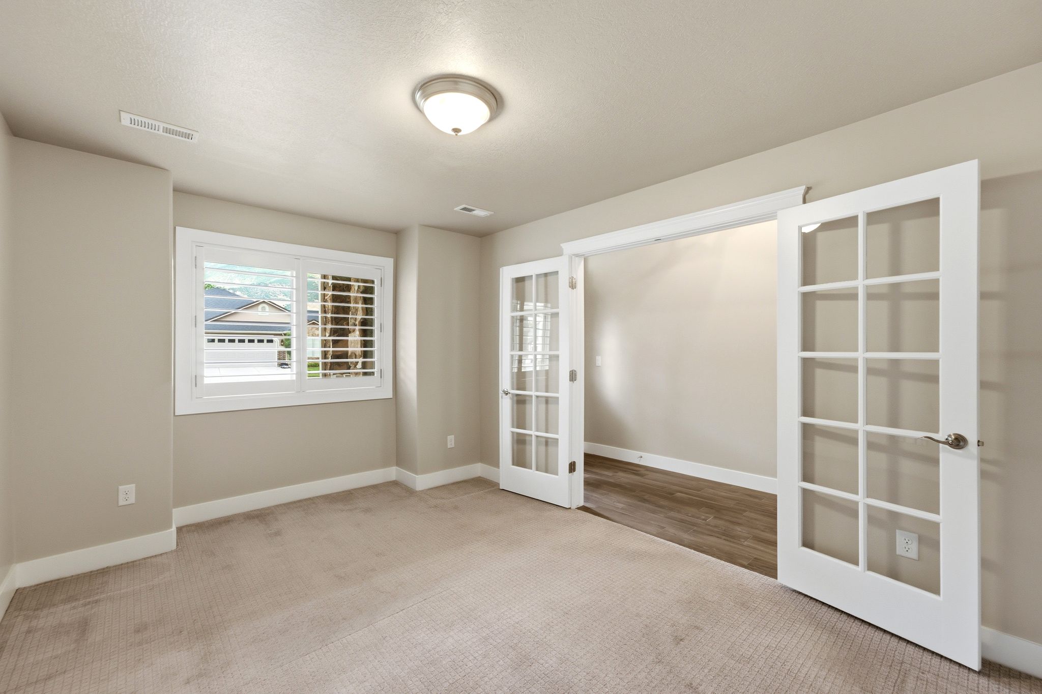 Carpeted room with a ceiling-mounted light fixture, white window trim, plantation shutters, and double French doors opening to an adjacent room with wood-finish flooring