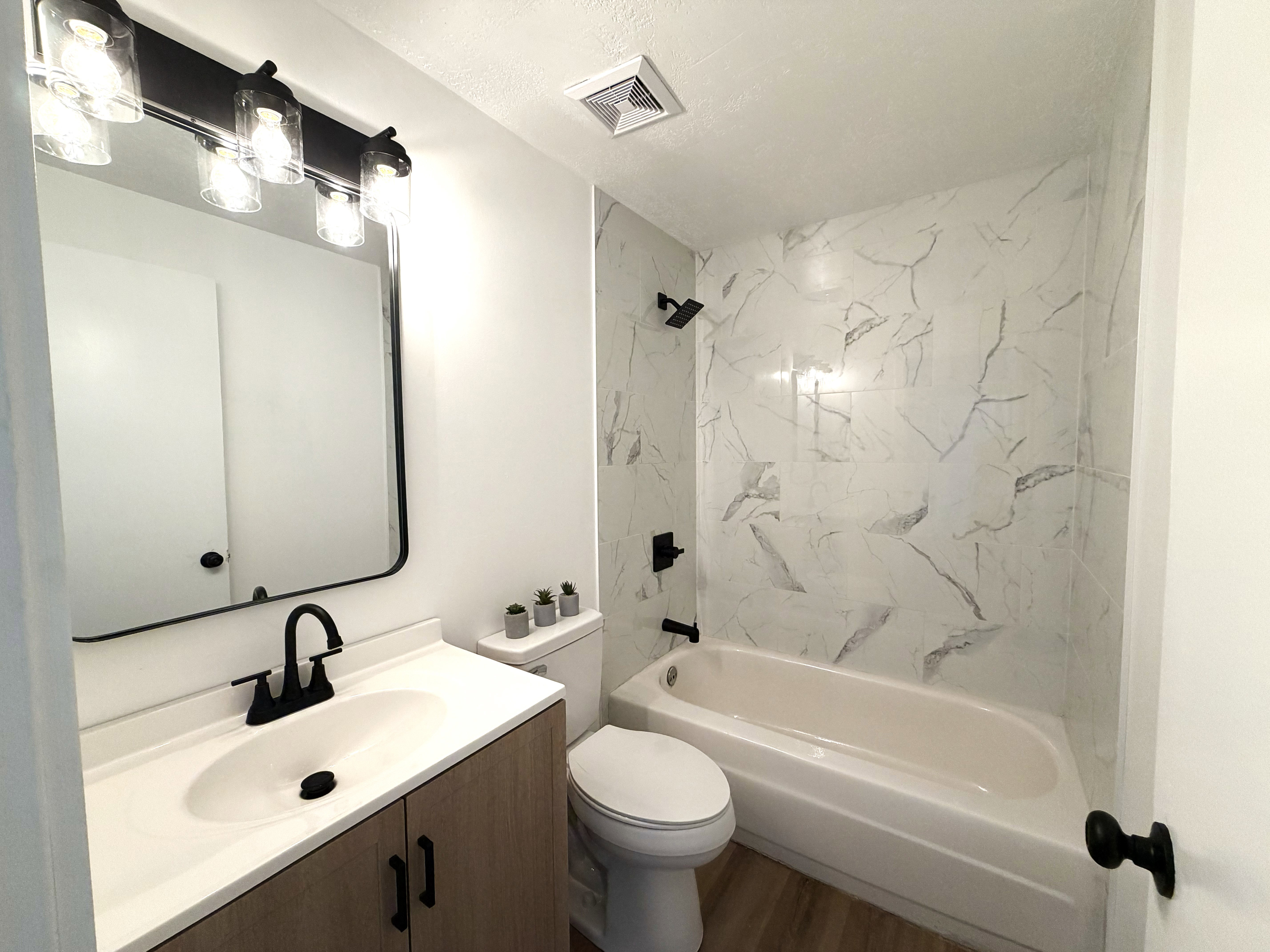 Bathroom featuring a wood-finish vanity with an integrated sink, matte black fixtures, a framed rectangular mirror, and a tub/shower combination with a white and gray tiled surround