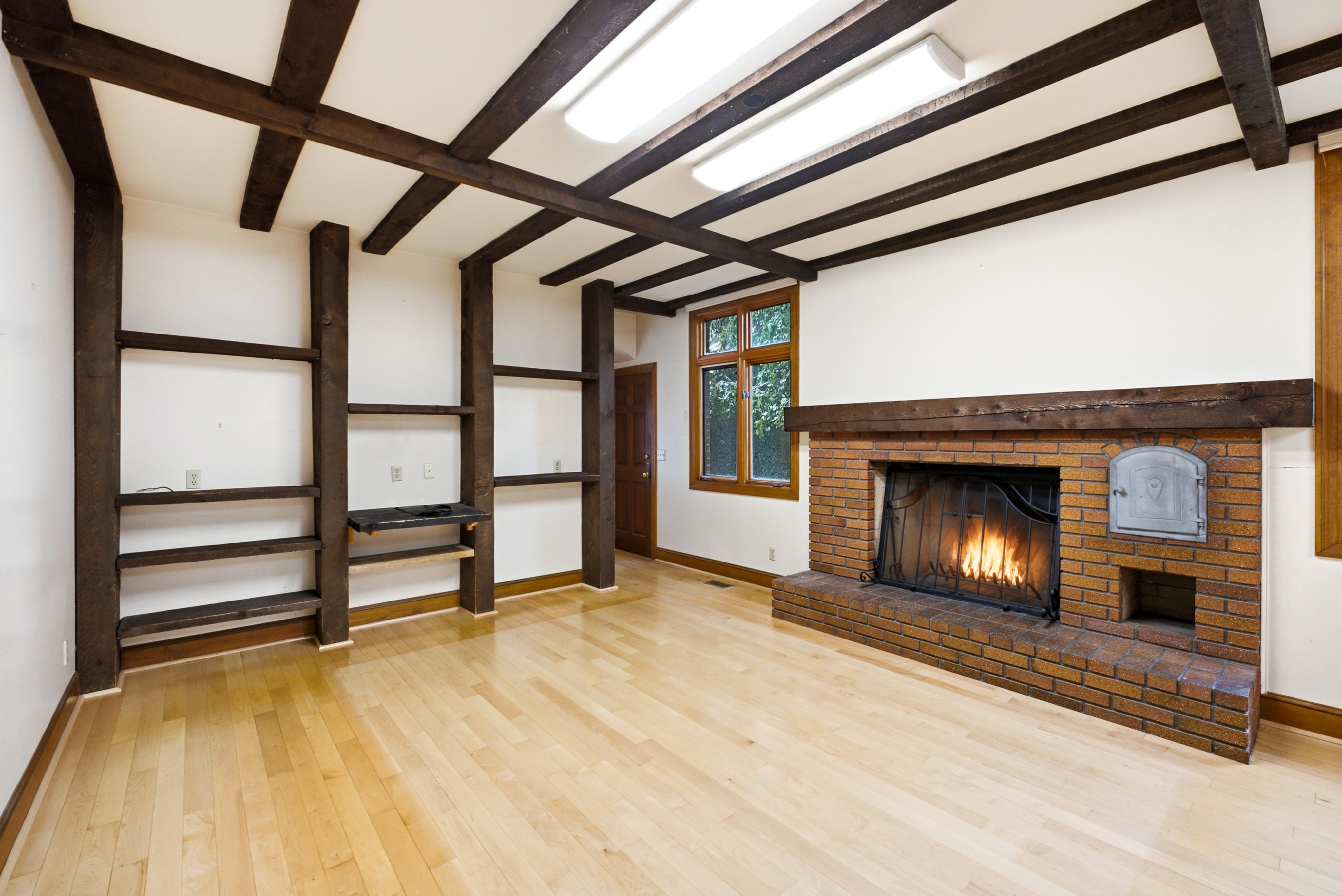 Living area featuring a brick fireplace with a wood-finish mantel, exposed wood ceiling beams, built-in shelving, wood-finish flooring, and a wood-frame window