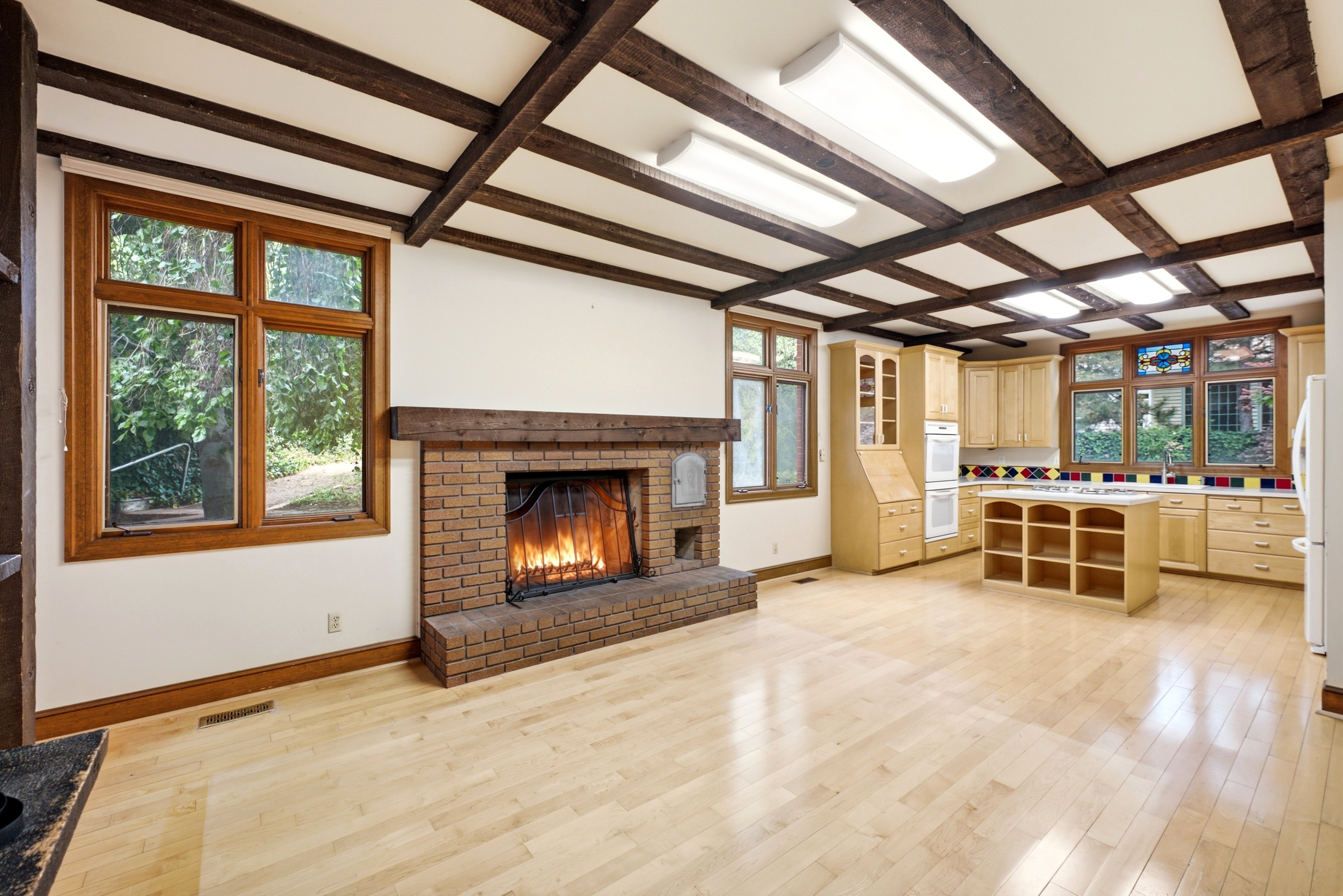 Open-concept room featuring a brick fireplace with a wood mantle, exposed wood beam ceiling, wood-finish flooring, light wood cabinetry, and multiple windows