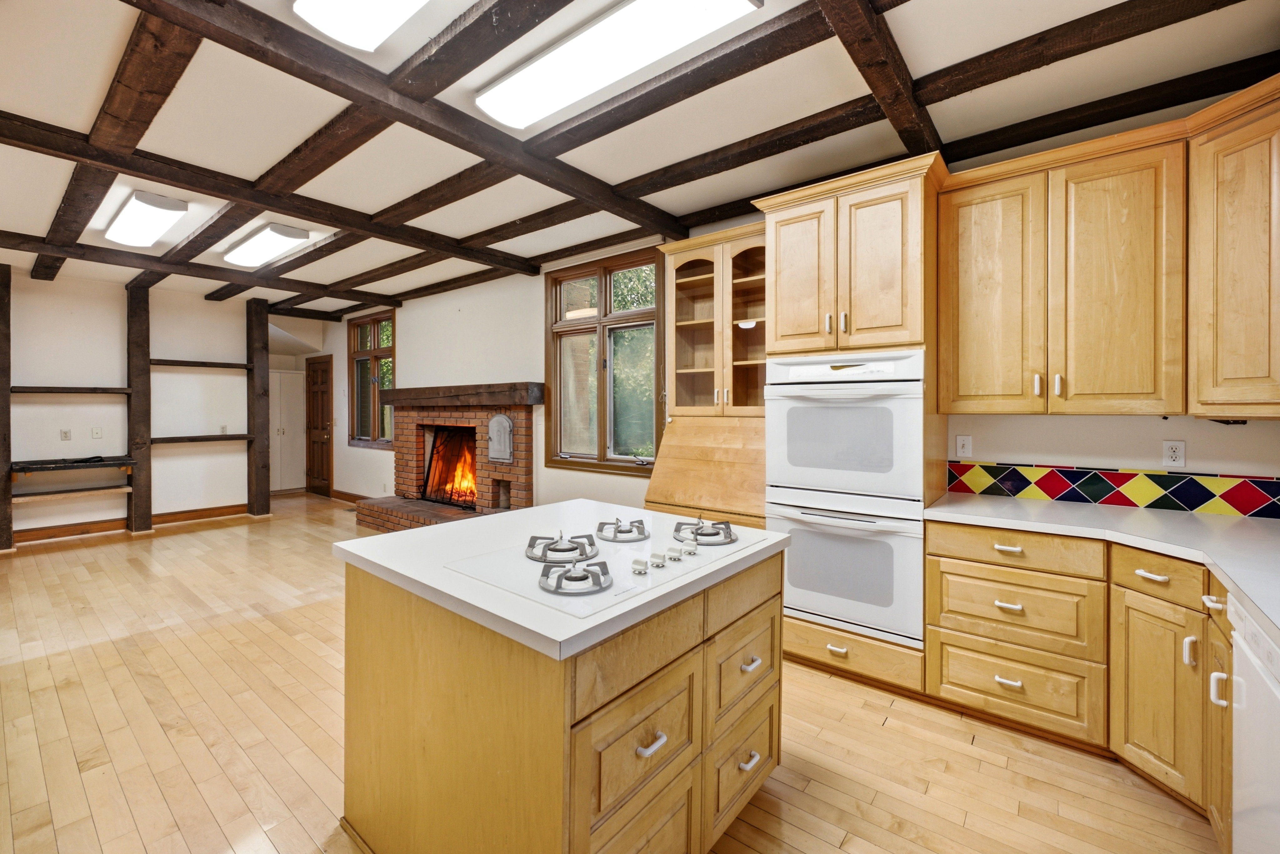 Open-concept kitchen and living area featuring a brick fireplace, exposed wood beam ceiling with recessed lighting, and light wood-finish flooring