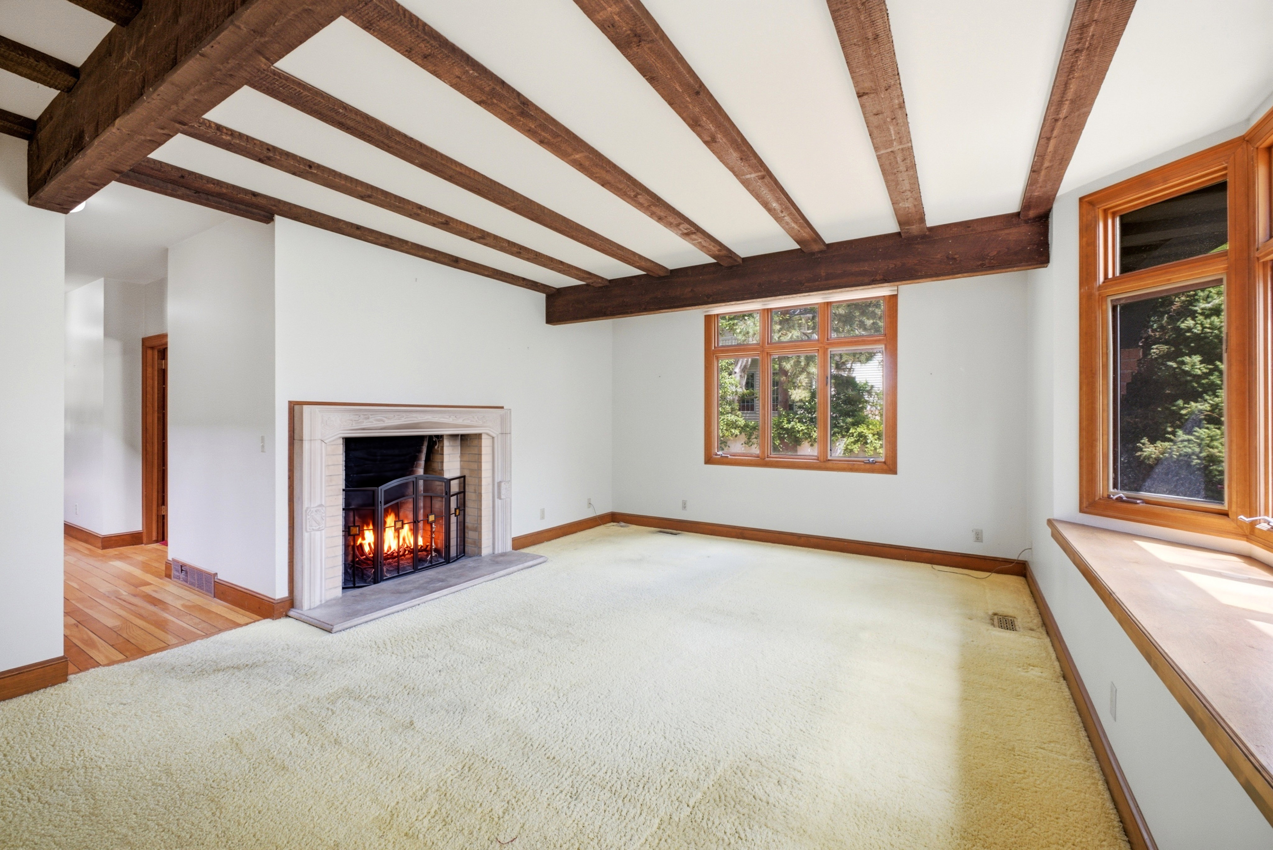 Living area featuring exposed wood ceiling beams, a stone-surround fireplace with a hearth, multiple wood-framed windows, and light-colored carpeting