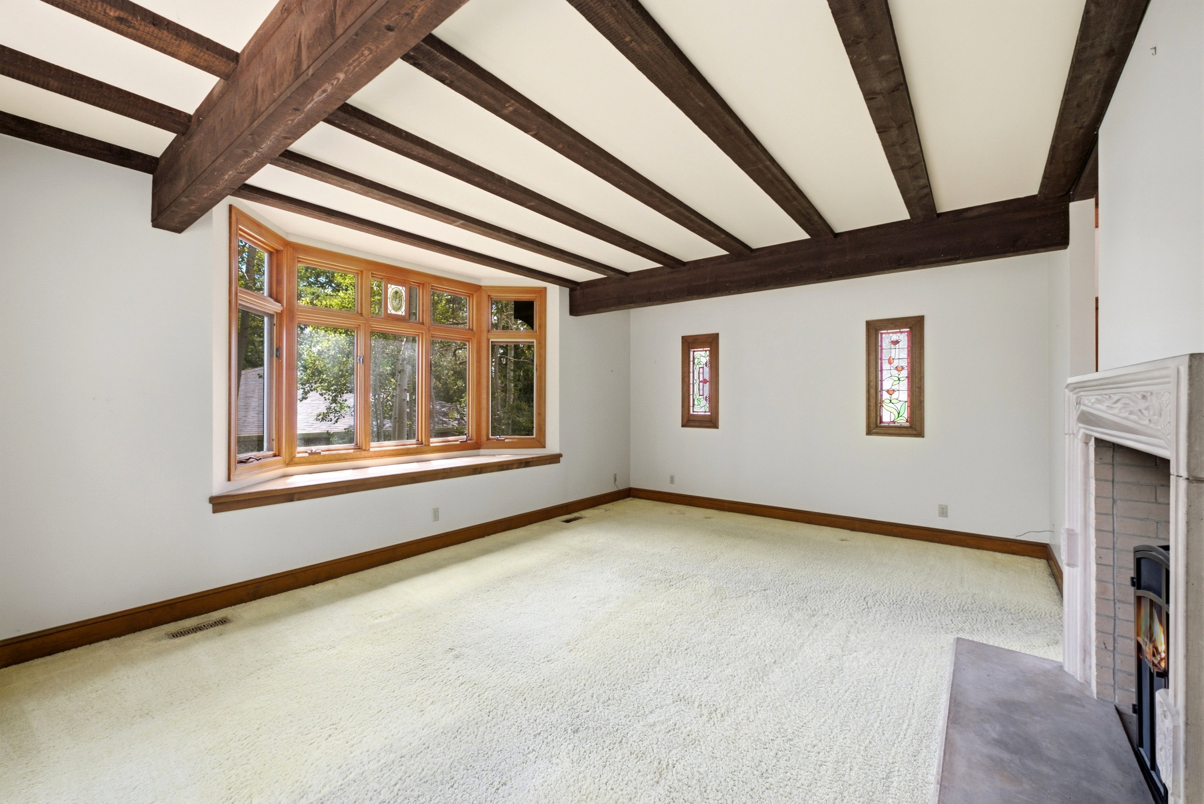 Living space featuring a wood-framed bay window, exposed wood ceiling beams, white carpeting, and a fireplace with a decorative mantel