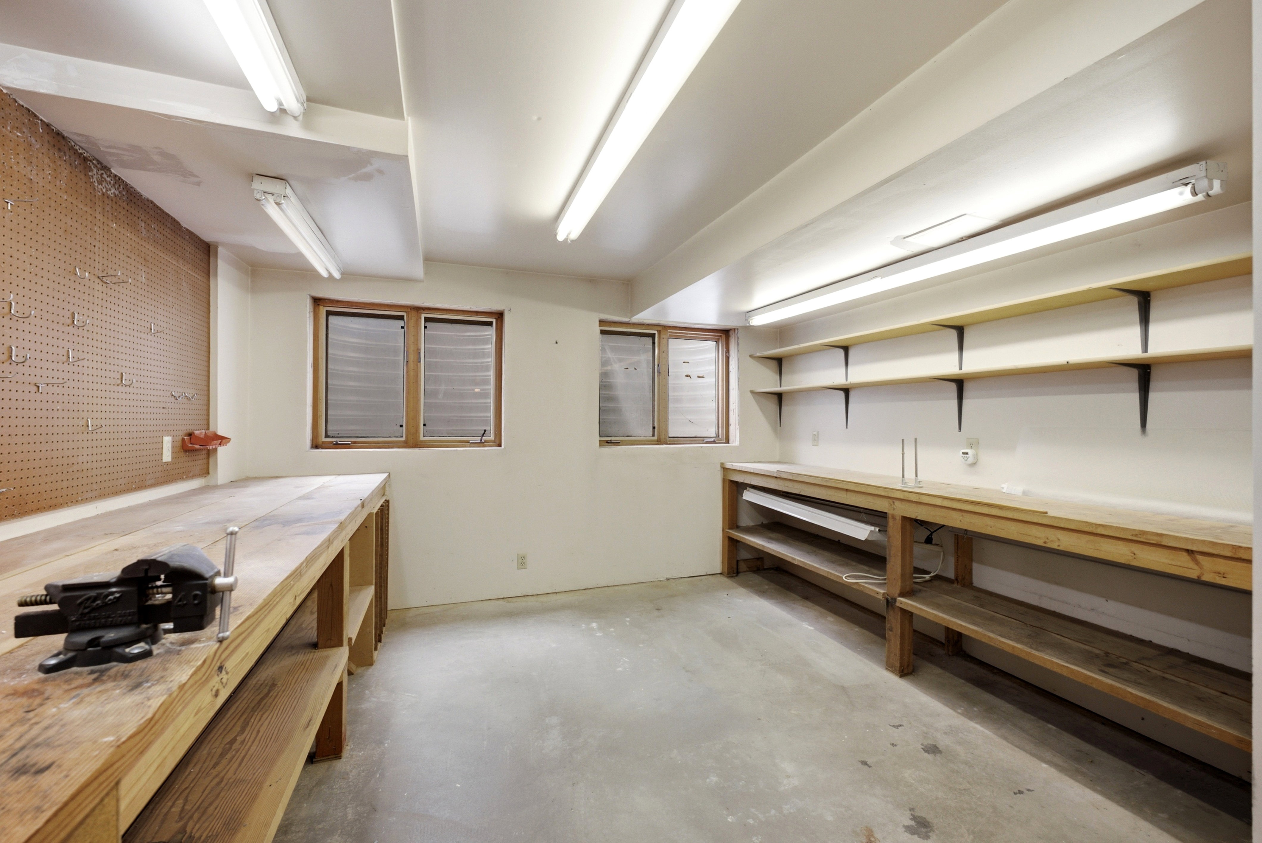 Utility room featuring concrete flooring, built-in wooden workbenches, wall-mounted shelving, pegboard storage, and multiple fluorescent light fixtures