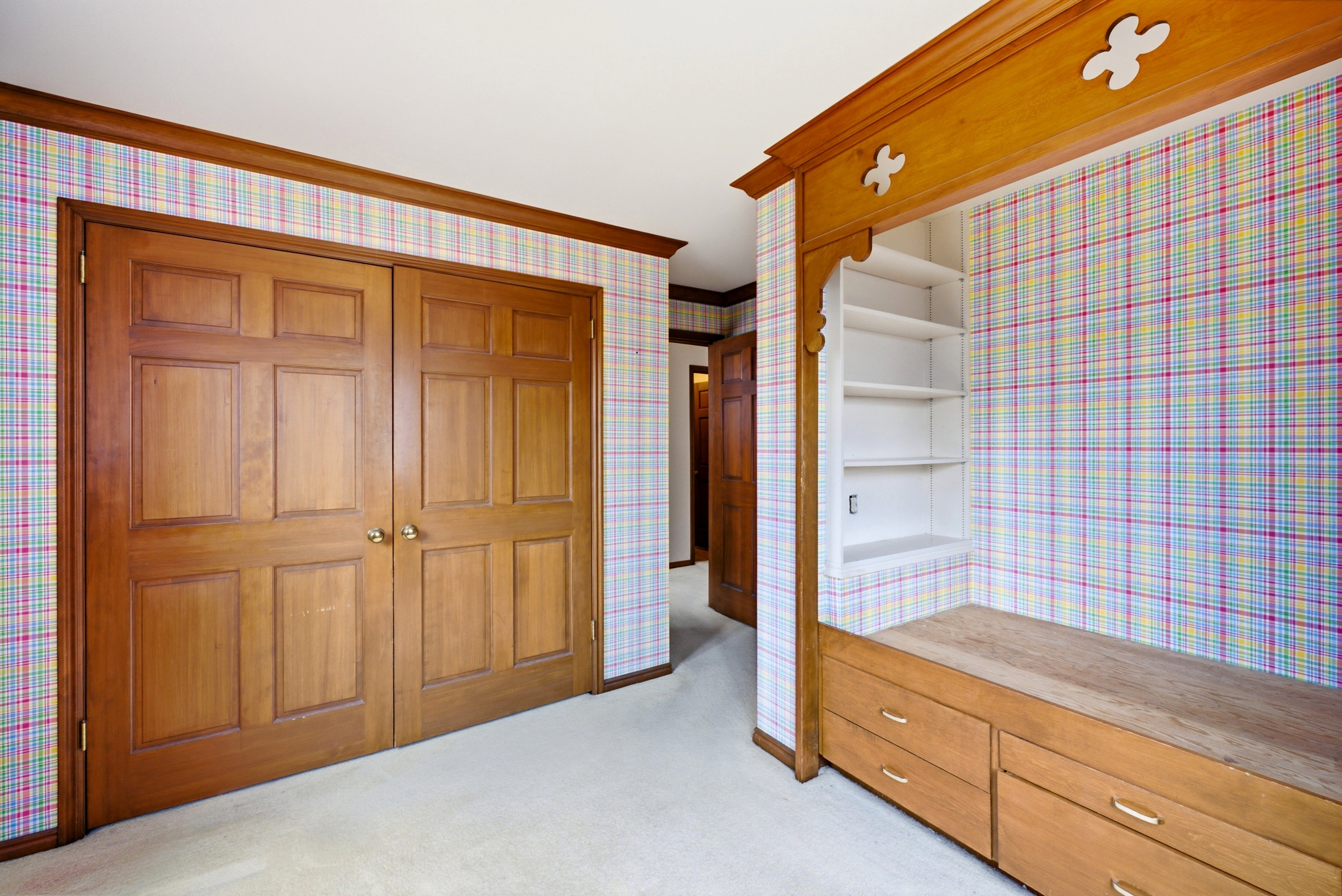 Room featuring a built-in storage unit with shelves and drawers, wood-finish bi-fold doors, wood-finish trim, and plaid wall covering