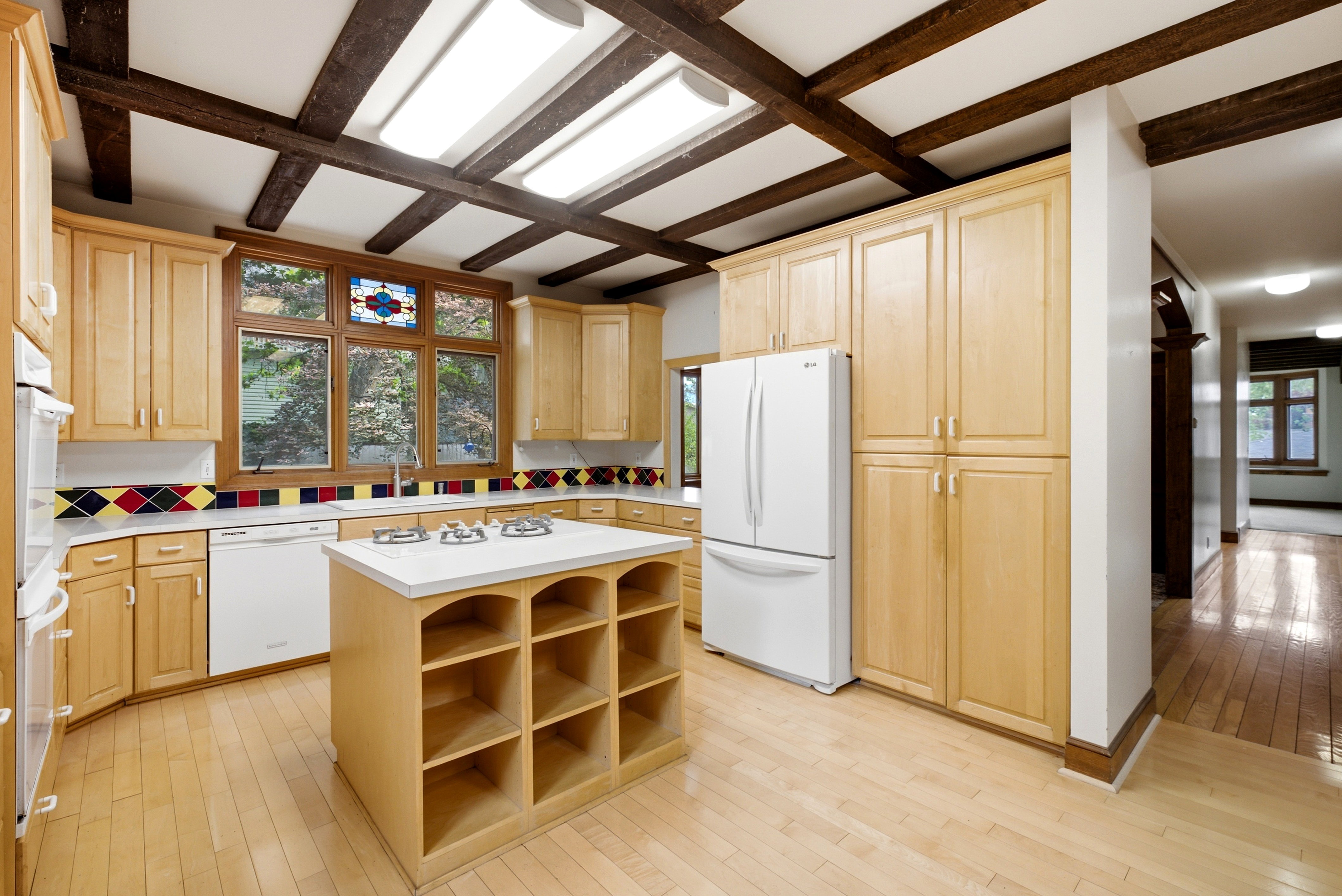 Kitchen featuring exposed ceiling beams, wood-finish flooring, light wood cabinetry, a central island with integrated cooktop, and a mosaic tile backsplash