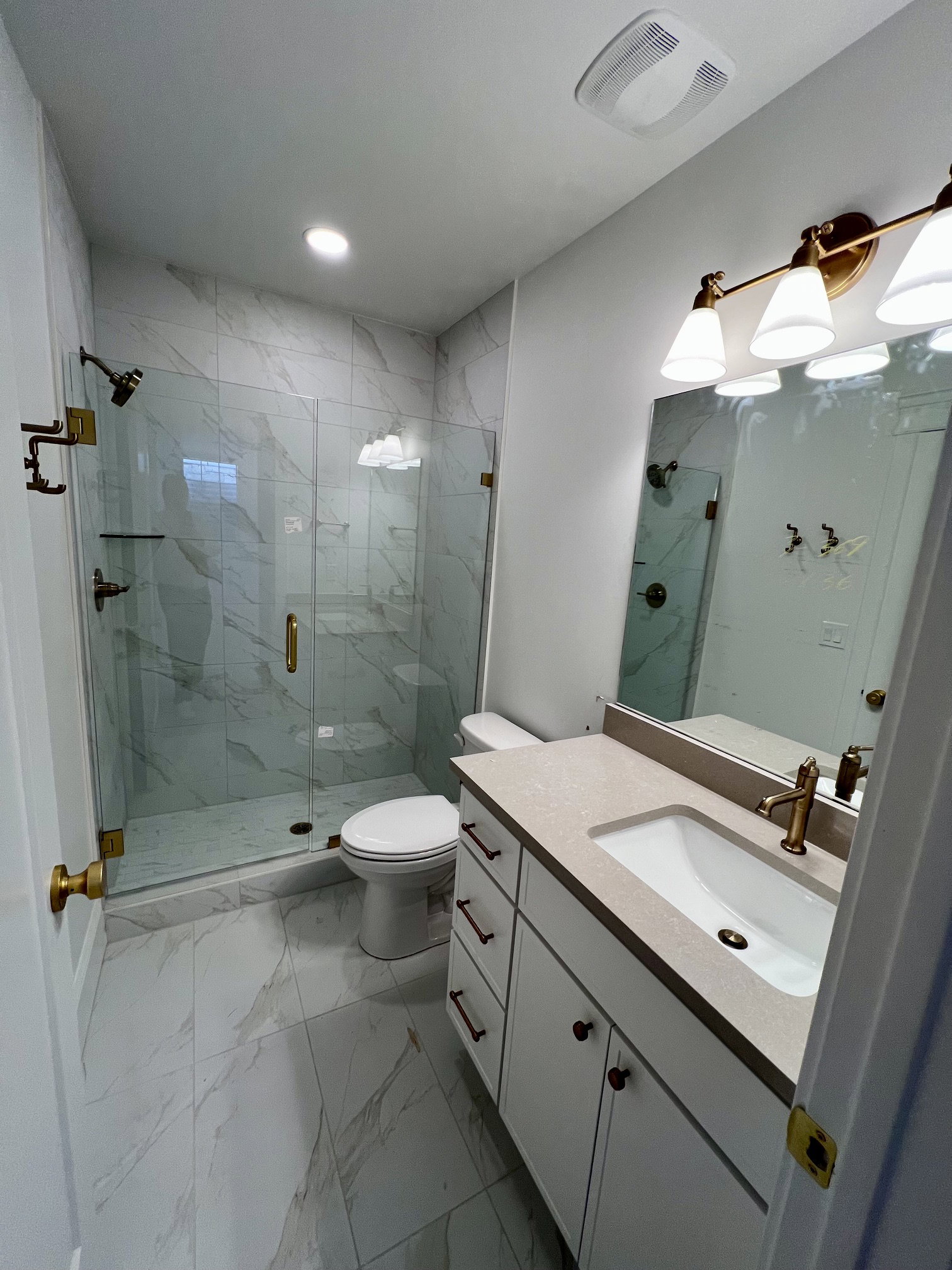 Contemporary bathroom featuring a walk-in shower with clear glass enclosure and gold-toned hardware, white marble-look tile flooring and shower surround, a white vanity with slab countertop, and a rectangular undermount sink with gold-toned faucet