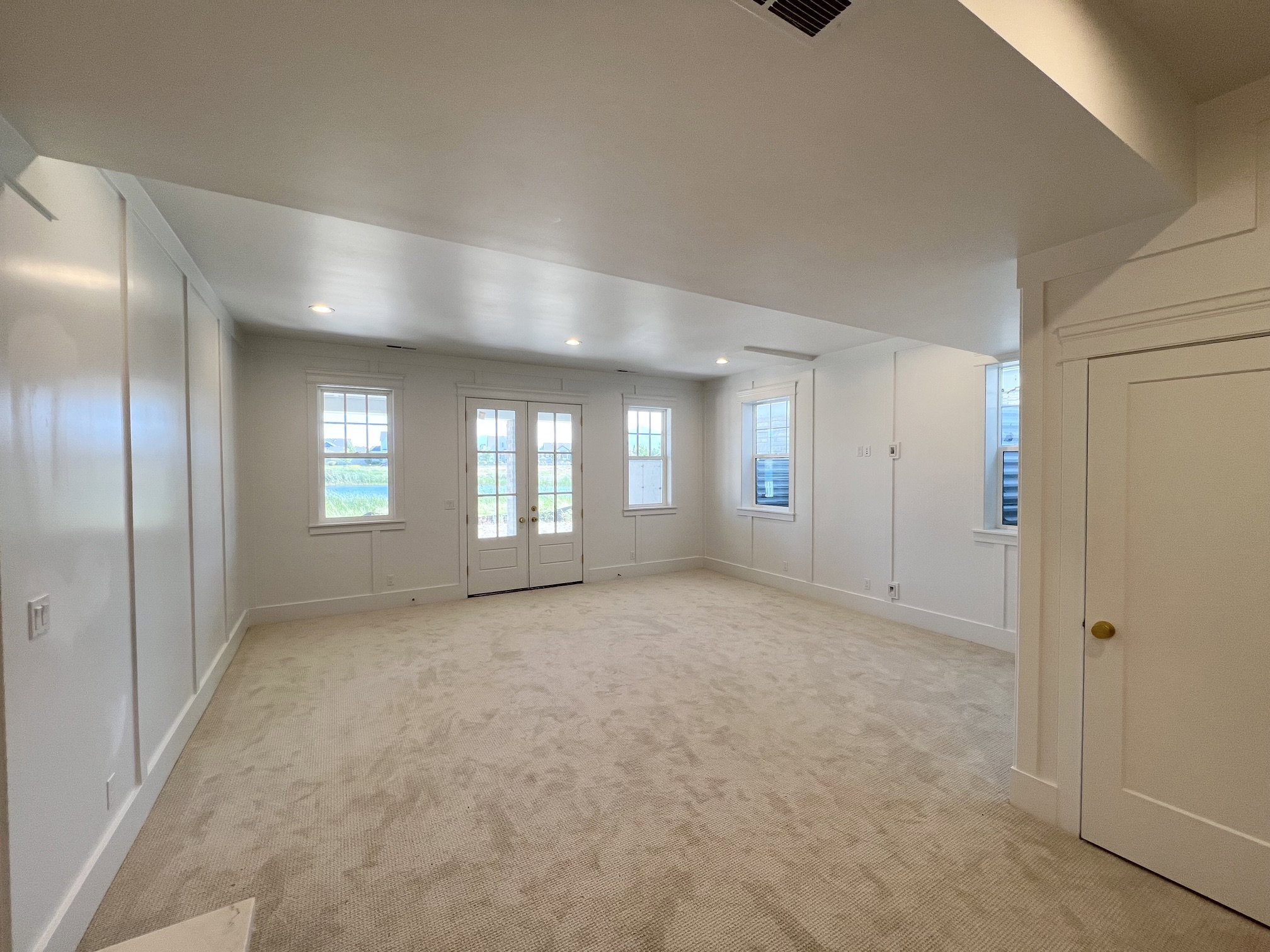 Spacious room featuring light beige carpeting, multiple windows with white trim, double French doors, recessed ceiling lighting, and crisp white wainscoting