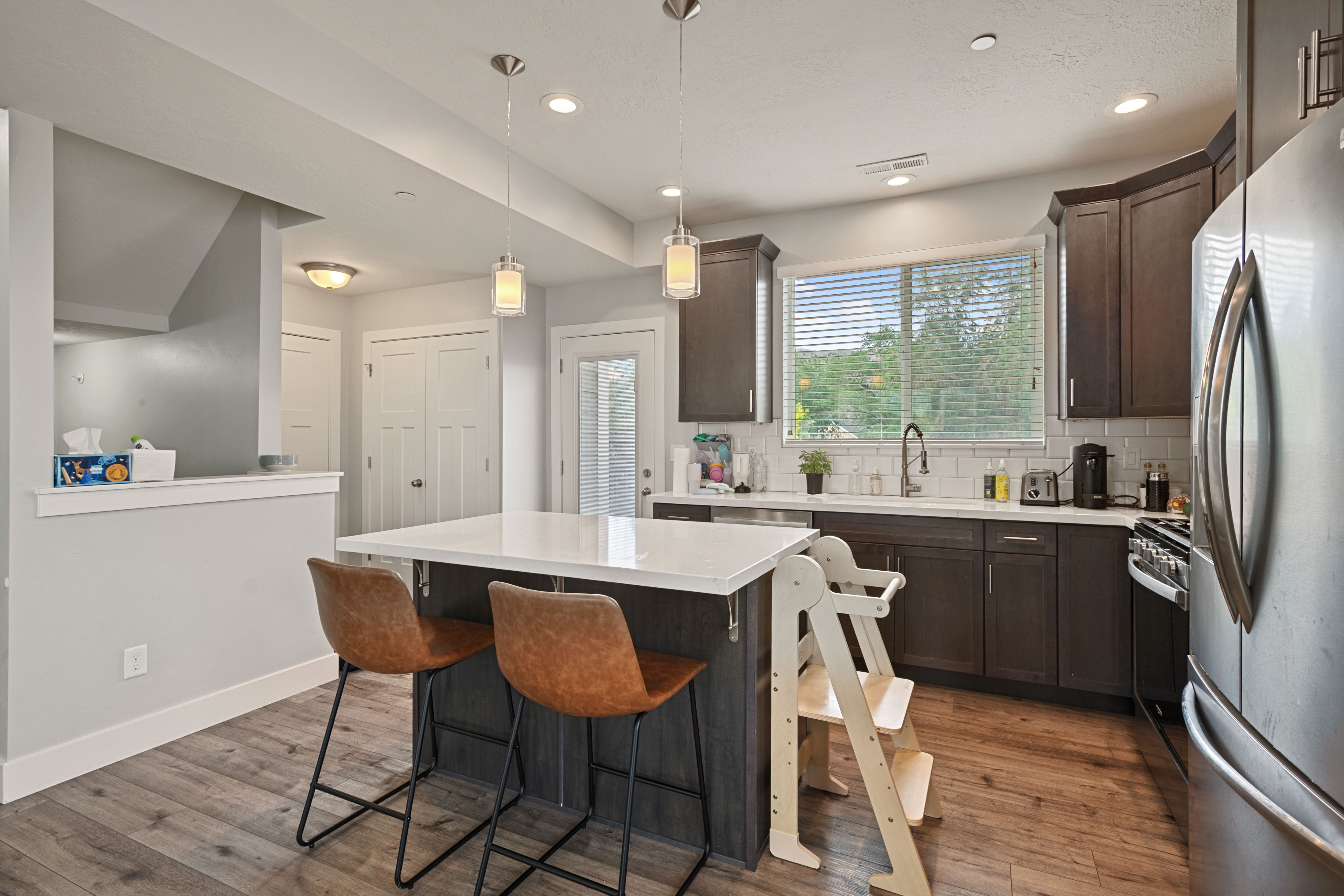 Modern kitchen featuring wood-finish flooring, dark wood cabinetry, white subway tile backsplash, and a central island with a white countertop and pendant lighting