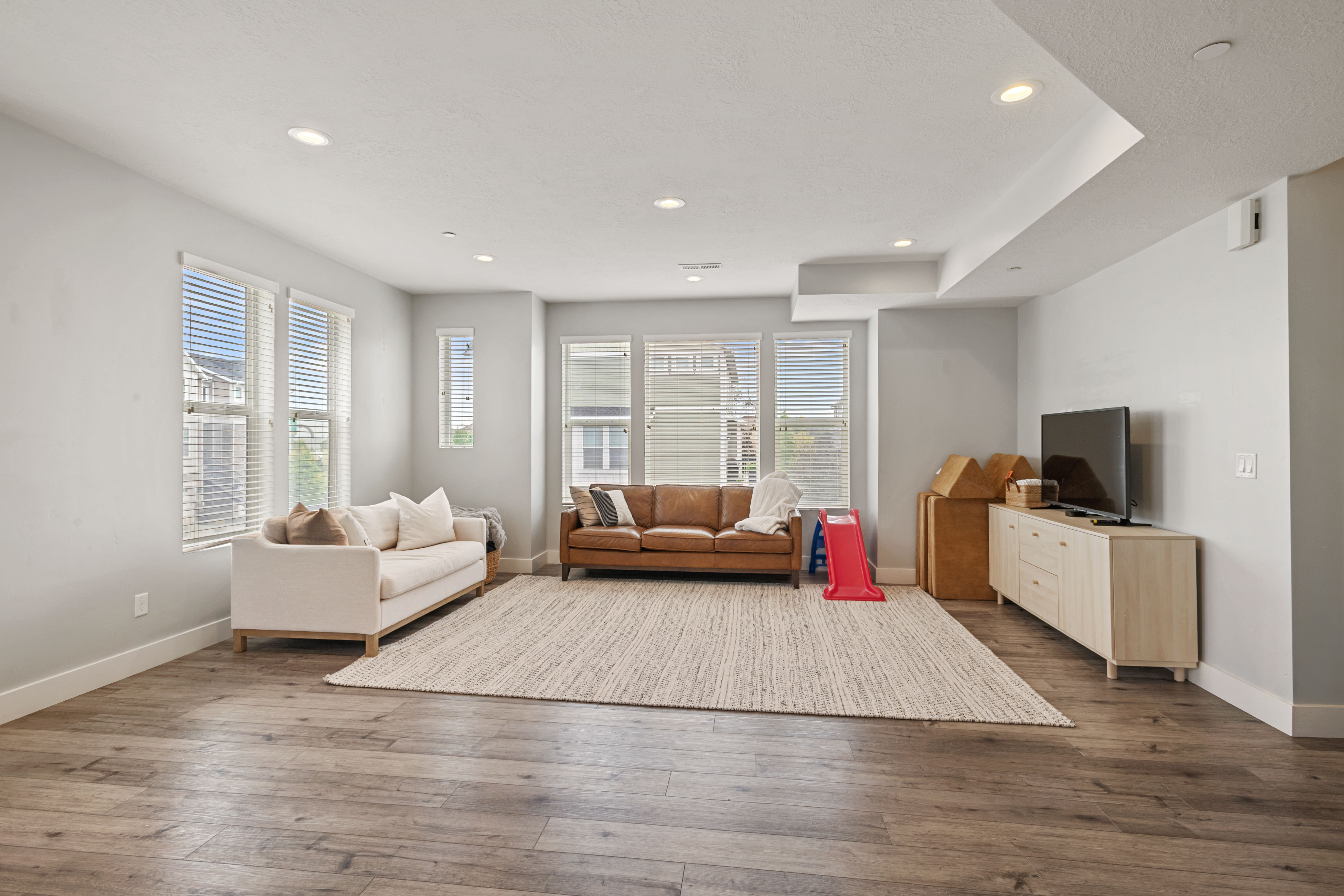 Spacious living area featuring wood-finish flooring, recessed lighting, multiple windows with blinds, and white baseboards