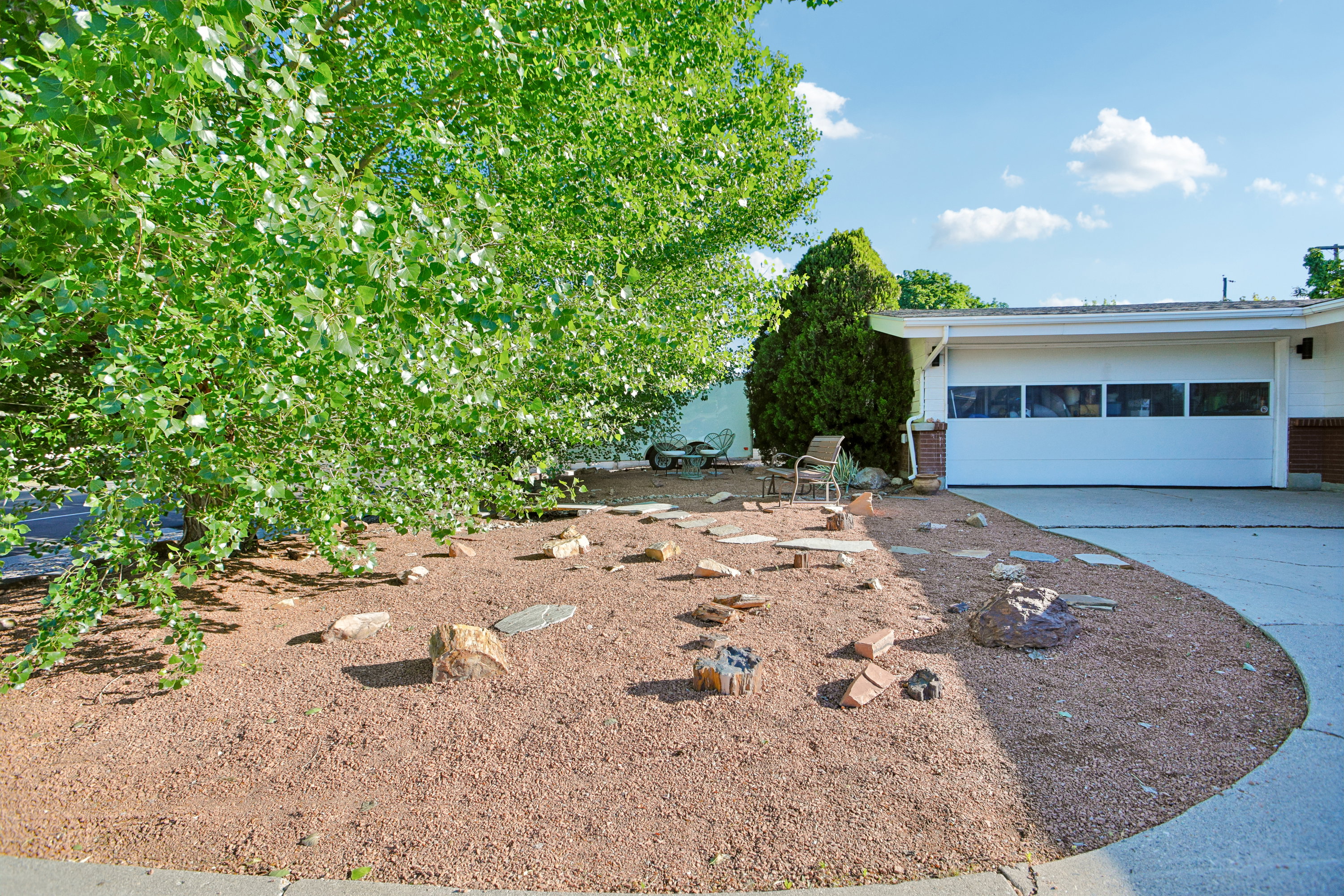 Front yard featuring extensive rock landscaping, mature deciduous trees, and a concrete driveway leading to a two-car garage with horizontal window inserts
