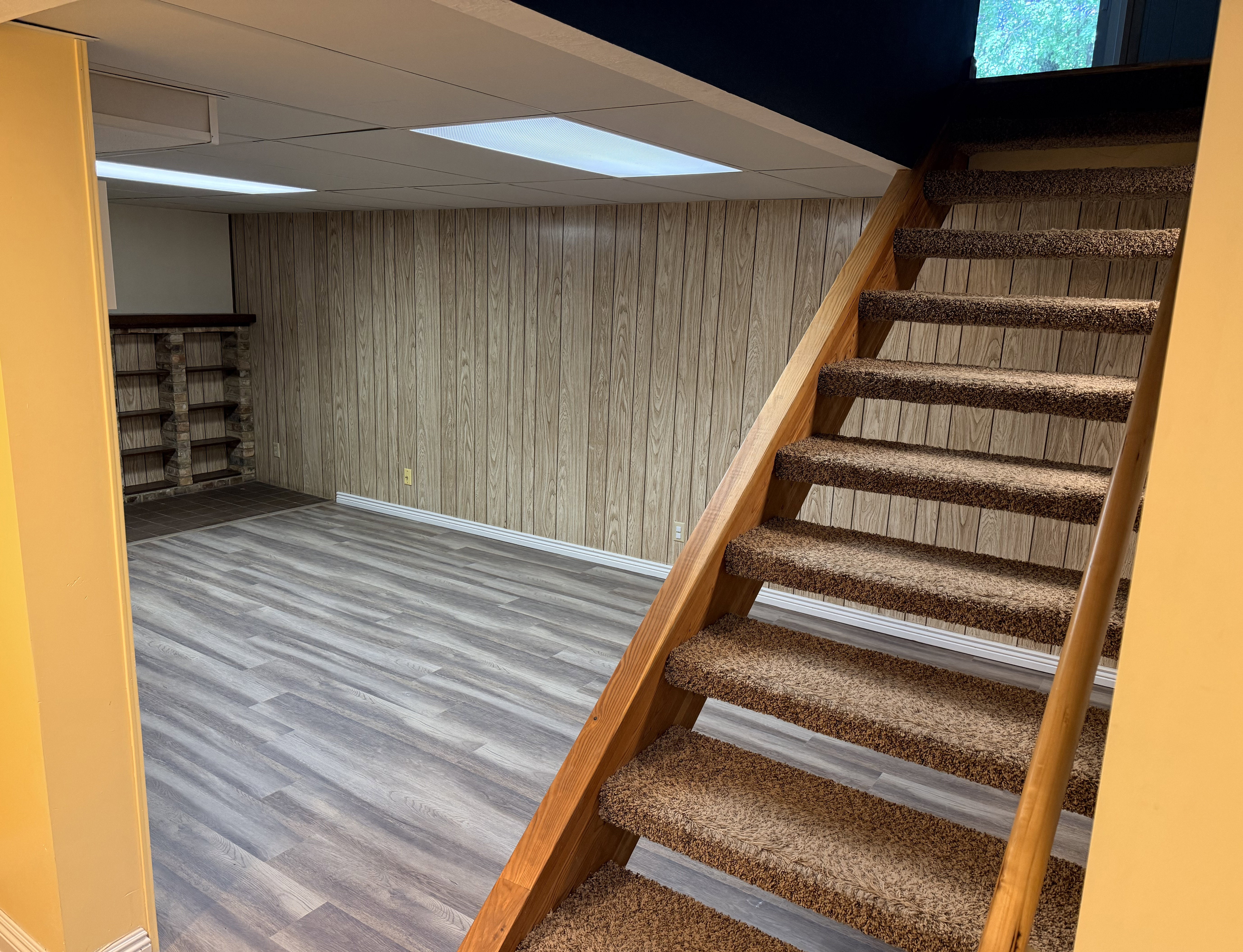 Carpeted staircase with wood handrail, leading to a finished room with wood-finish flooring, wood paneling, and built-in shelving