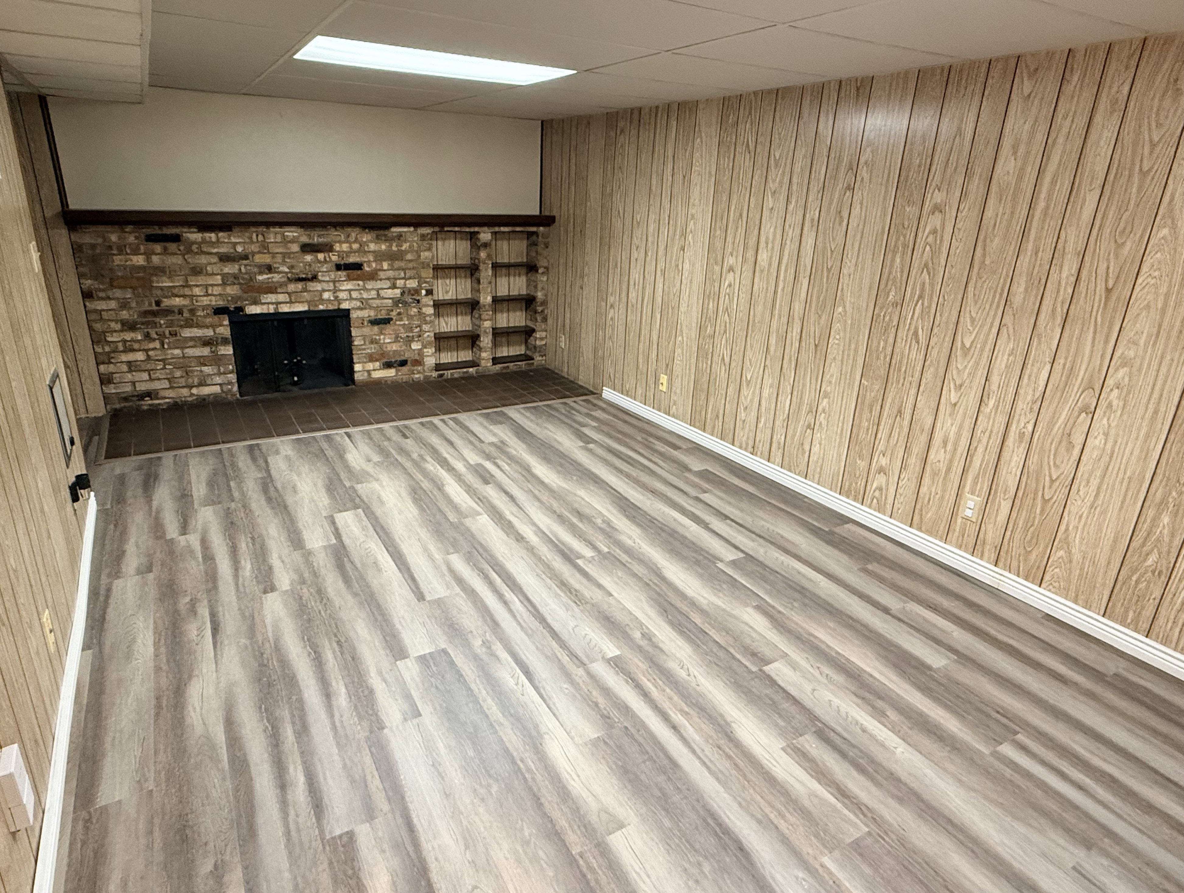 Spacious room featuring wood-finish paneling, a brick-front fireplace, built-in shelving, and light wood-finish flooring