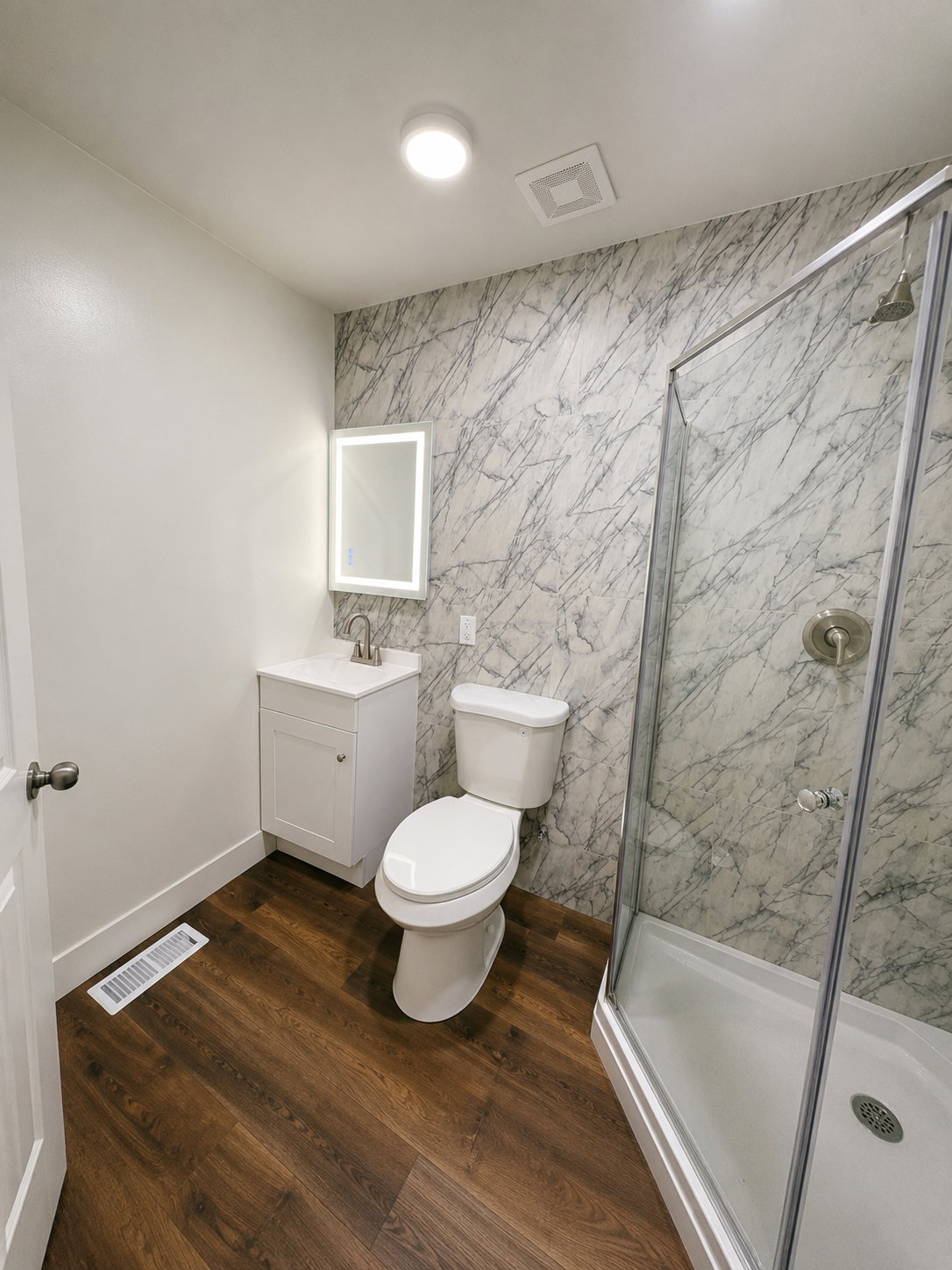 Full bathroom featuring wood-finish flooring, a white vanity with integrated sink, an illuminated mirror, a toilet, and a glass-enclosed corner shower with a brushed nickel shower head