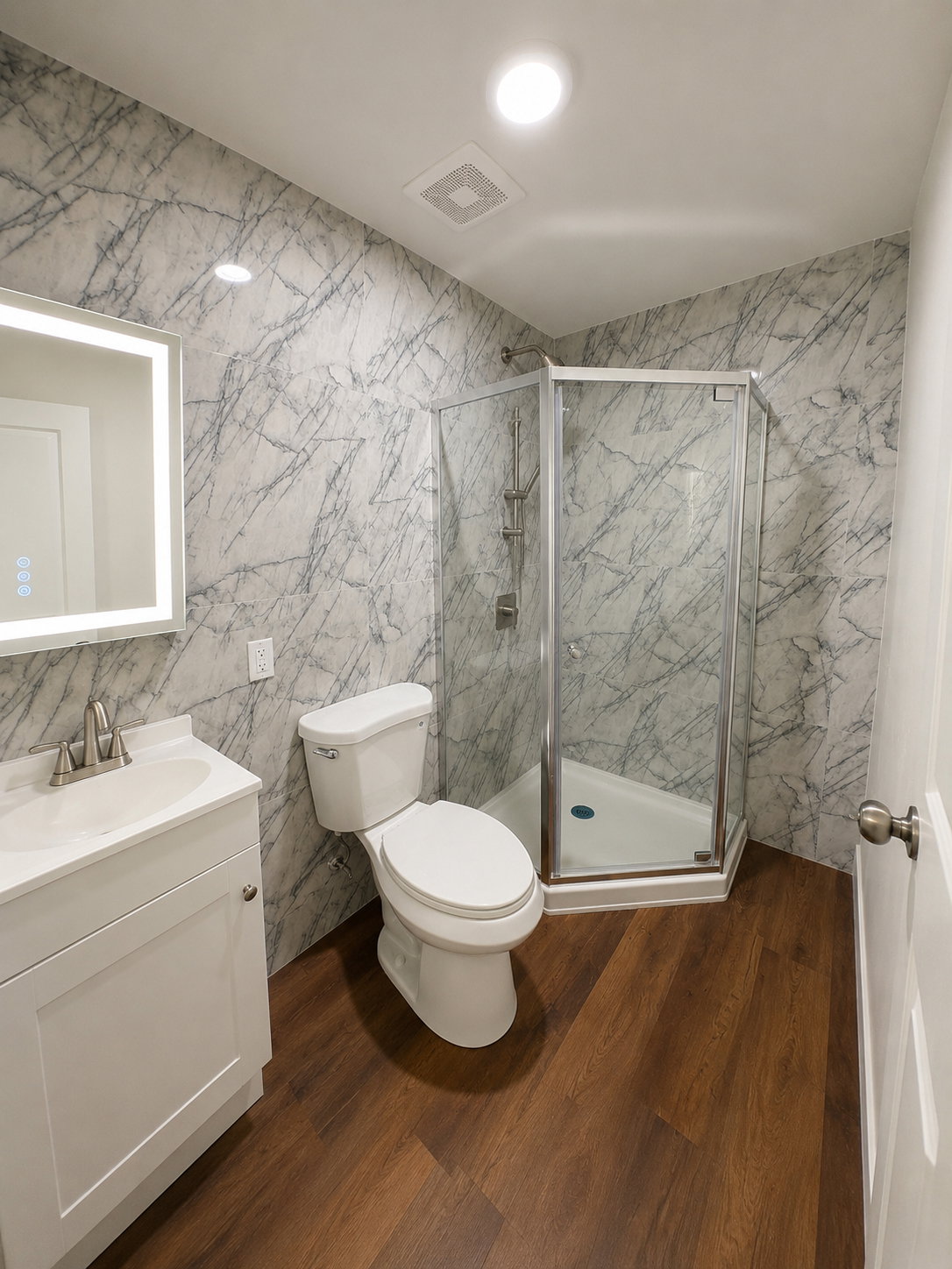 Modern bathroom featuring wood-finish flooring, marbled wall panels, white vanity with integrated sink, sleek backlit mirror, and a corner glass-enclosed shower