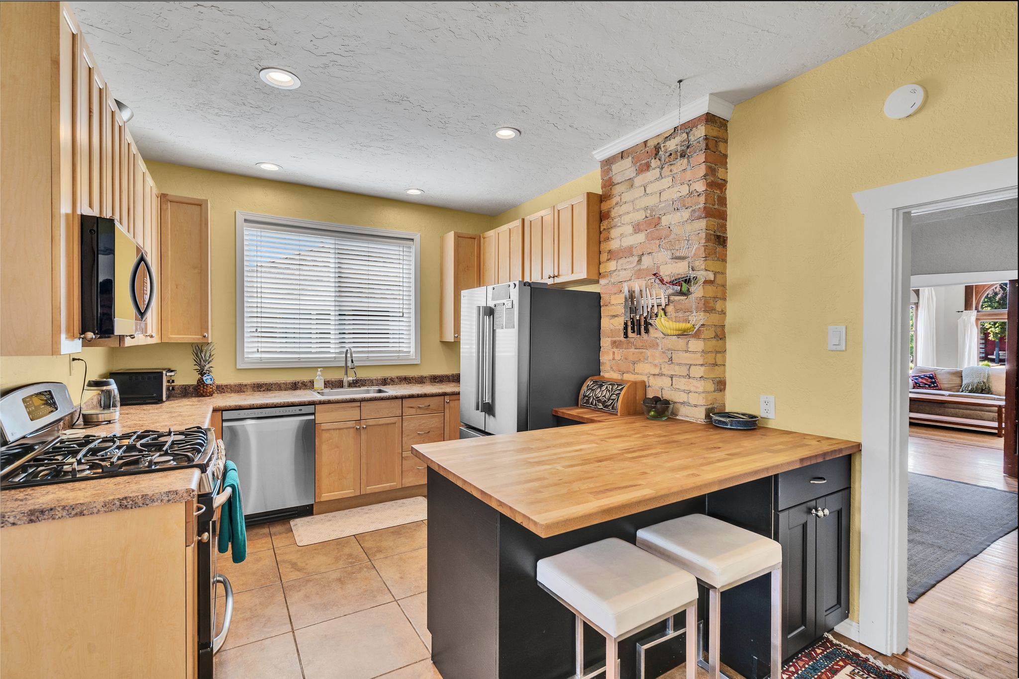 Kitchen featuring a original brick accent wall, light wood cabinetry, stainless steel appliances, a center island with a wood countertops, and tile flooring