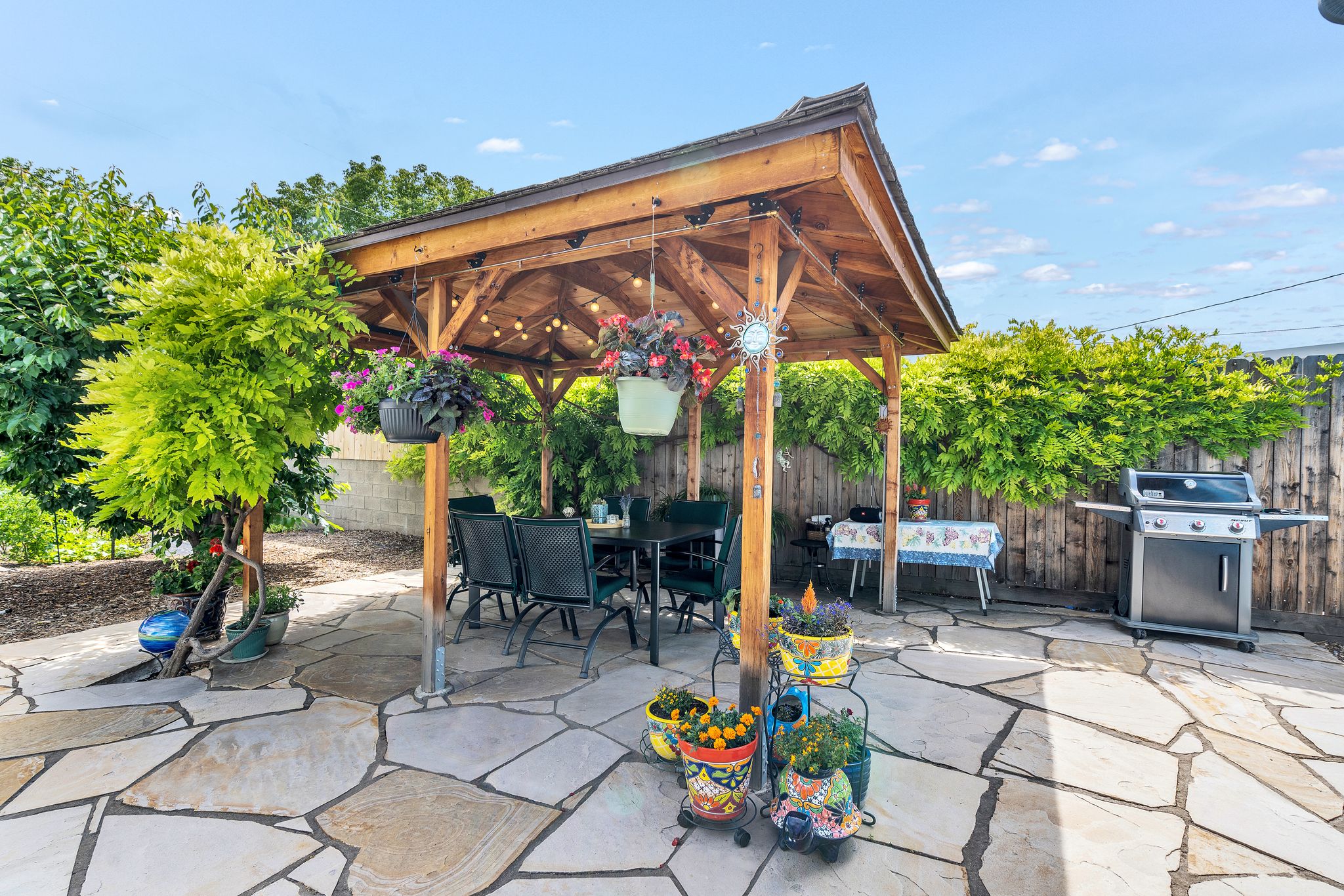 Spacious flagstone patio featuring a wooden pergola with string lighting, surrounded by mature trees and a privacy fence