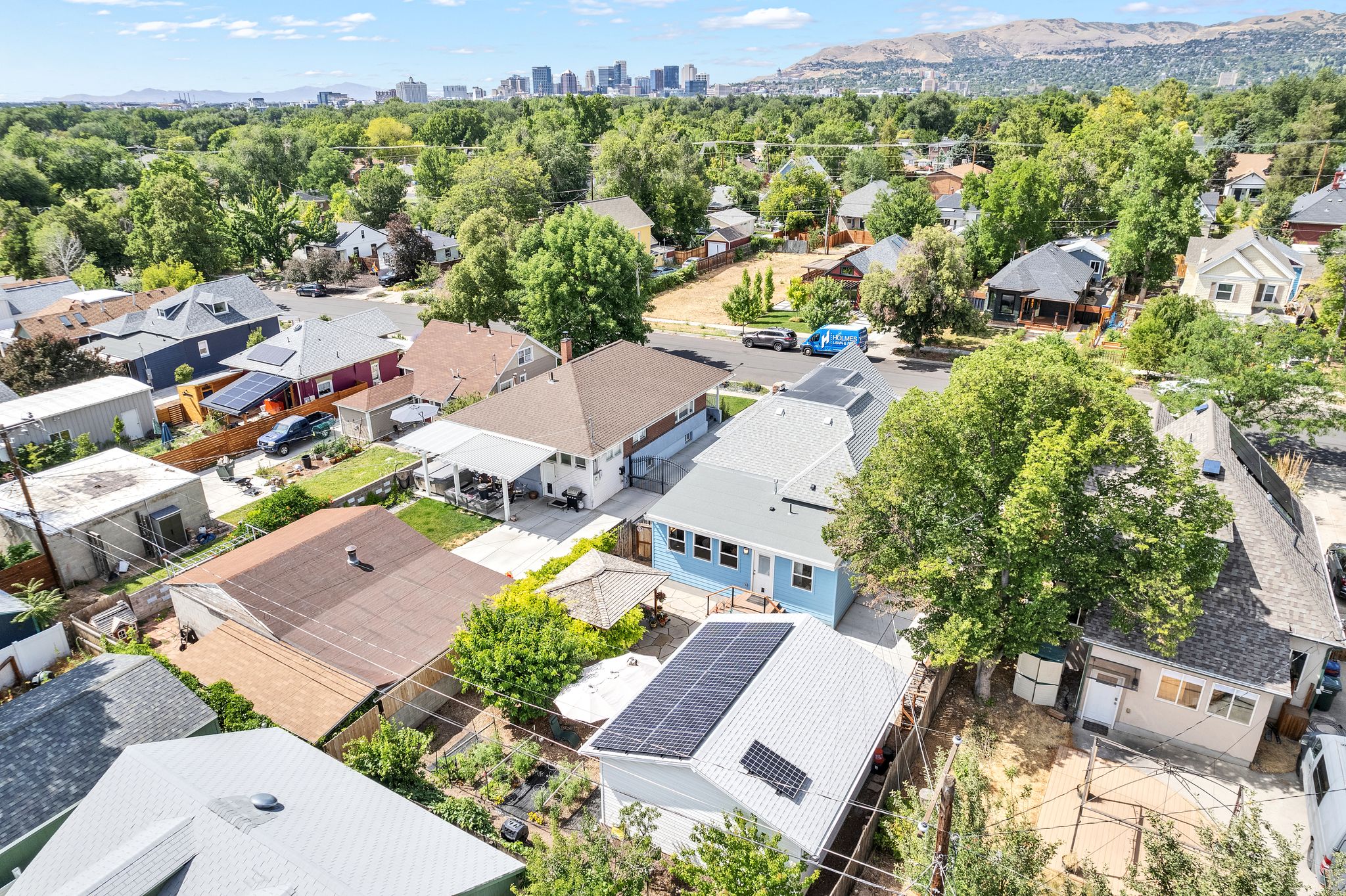Aerial perspective of a neighborhood with diverse residential structures, abundant mature trees, and a distant city skyline with mountains