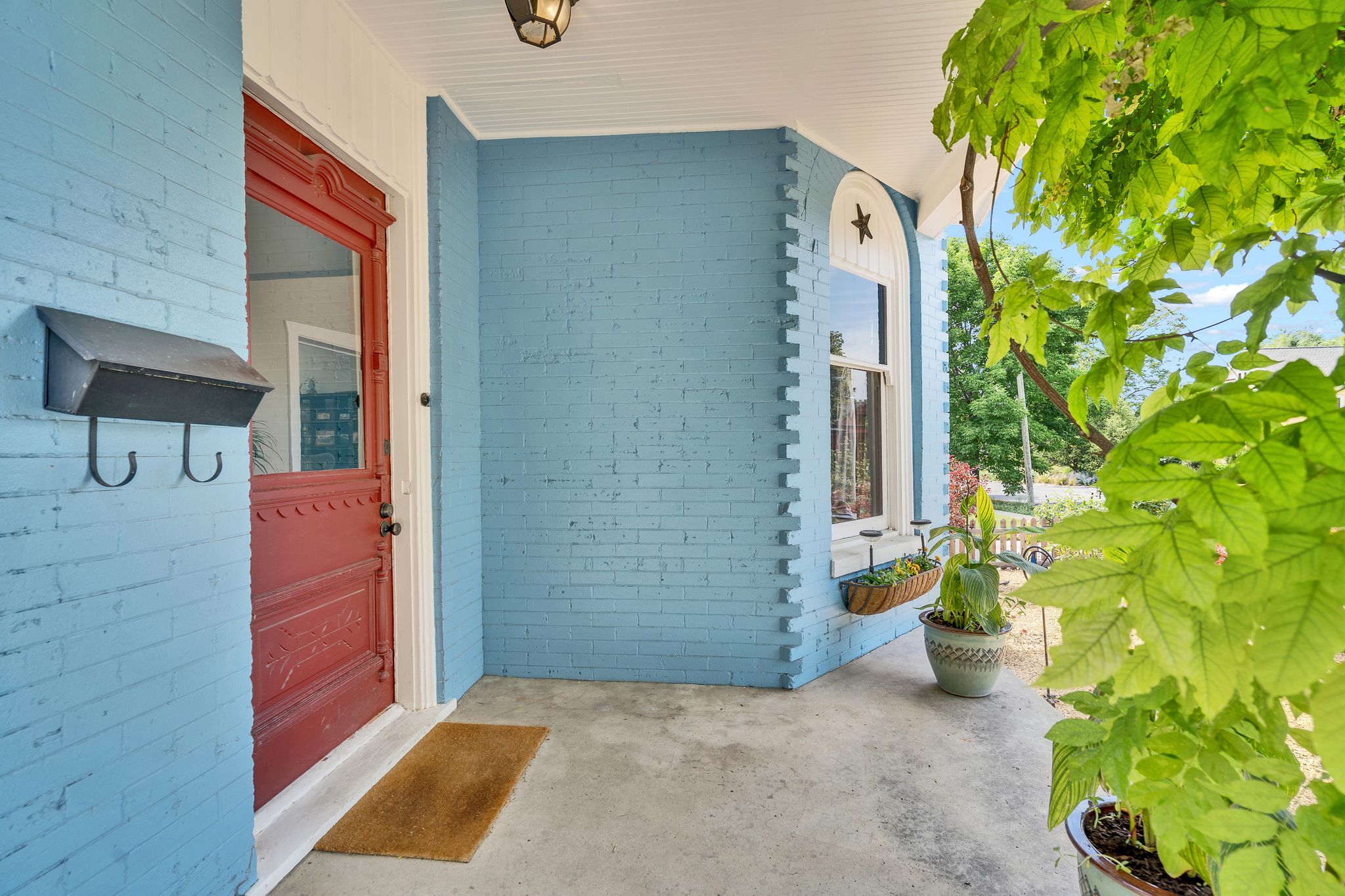 Covered porch entry featuring a decorative red door with glass panel, blue painted brick exterior, and a single arched window with white trim