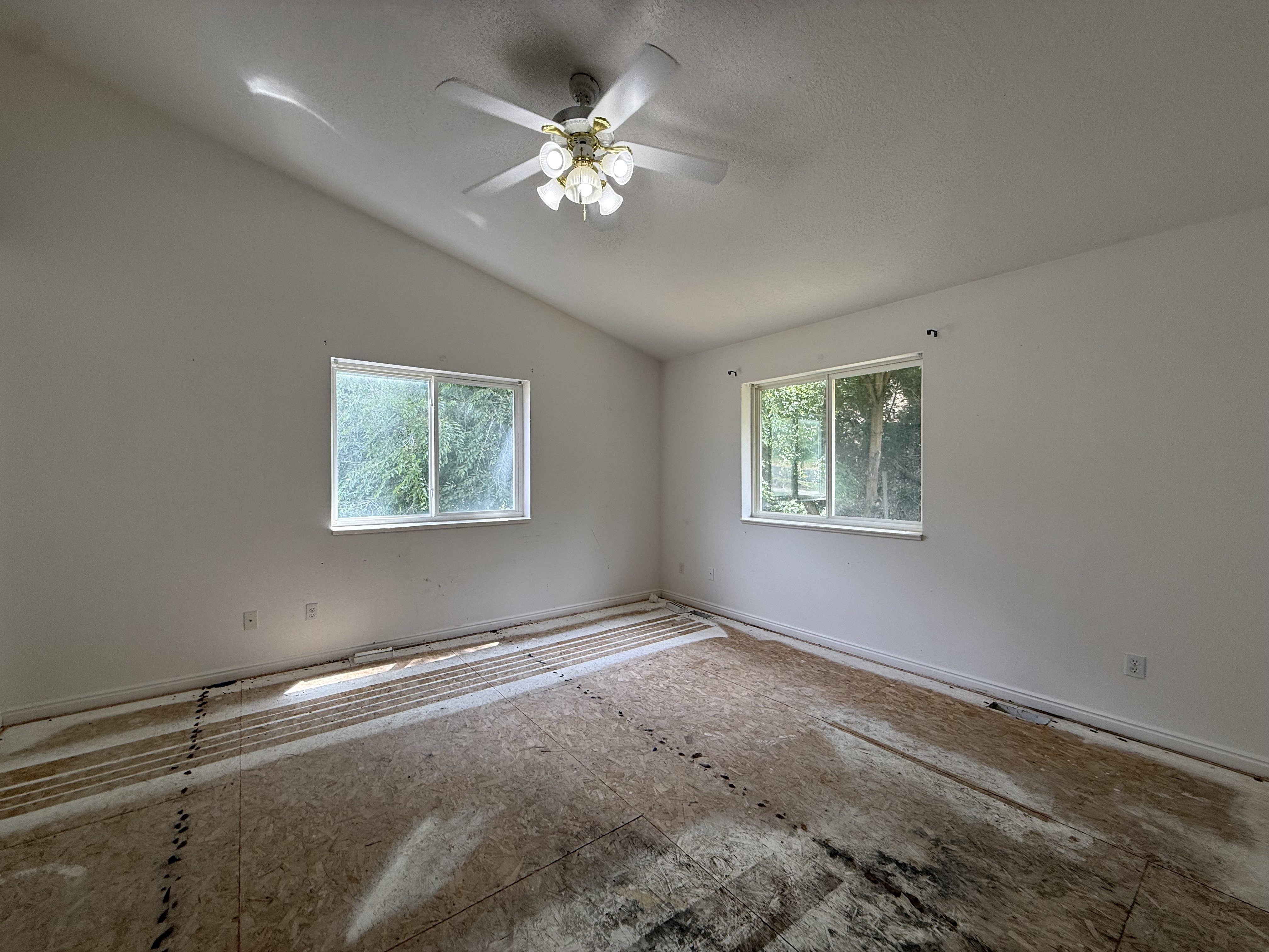 Vaulted ceiling room featuring two windows, a ceiling fan with light fixture, white walls, and subfloor