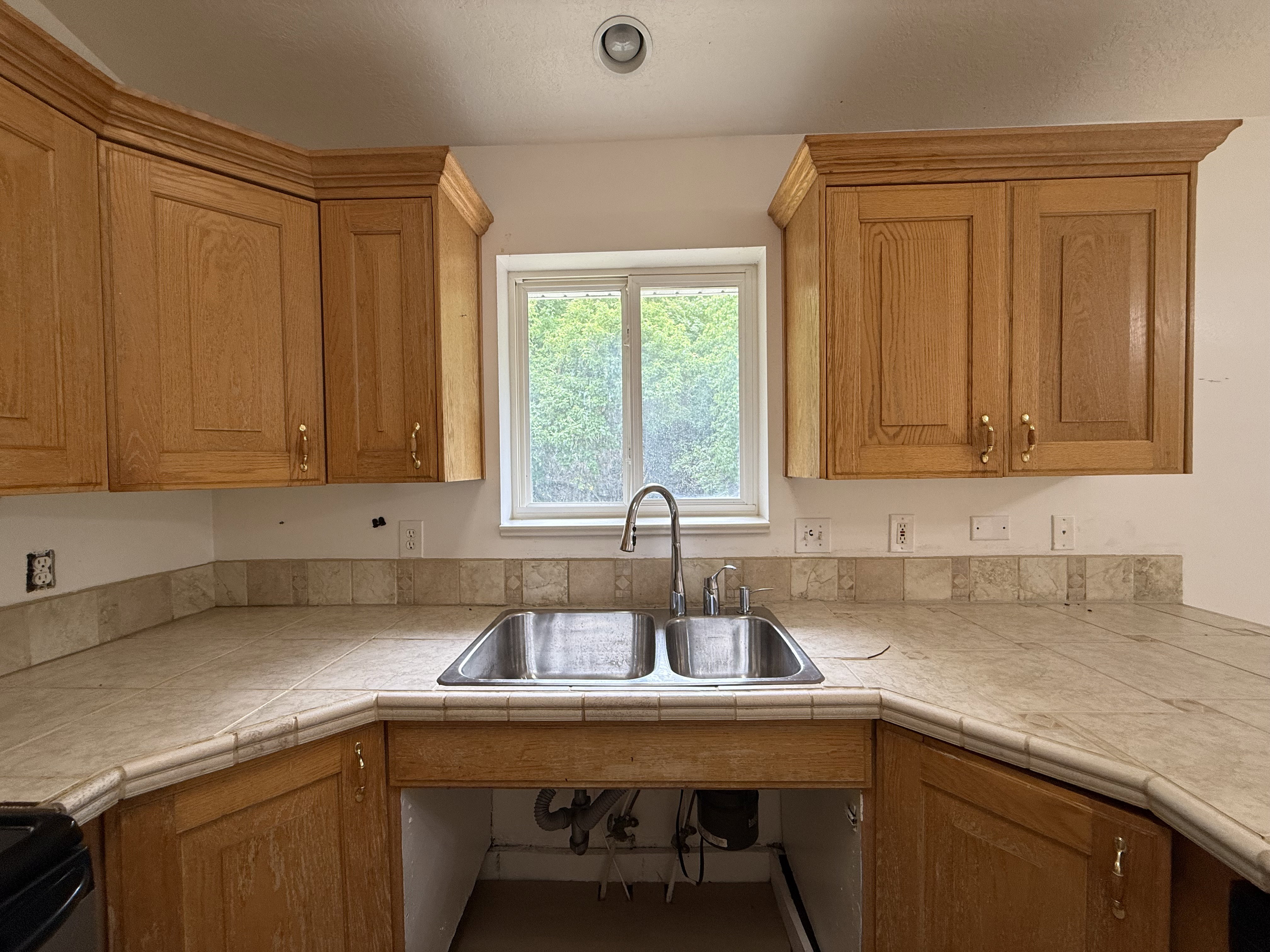 Kitchen featuring a double stainless steel sink, wood cabinetry, light-toned tile countertops, a matching tile backsplash, and a window above the sink