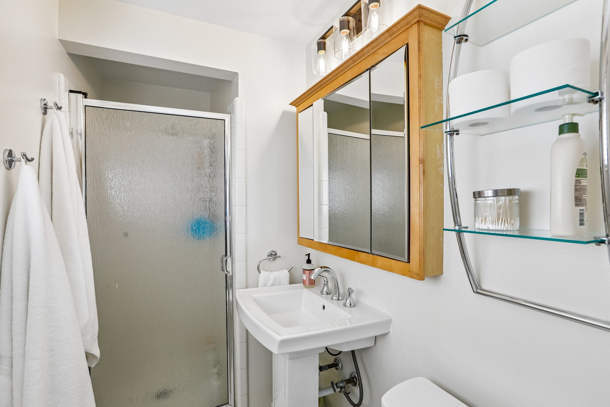 White pedestal sink with chrome faucet, complemented by a wood-finish medicine cabinet and overhead vanity lighting