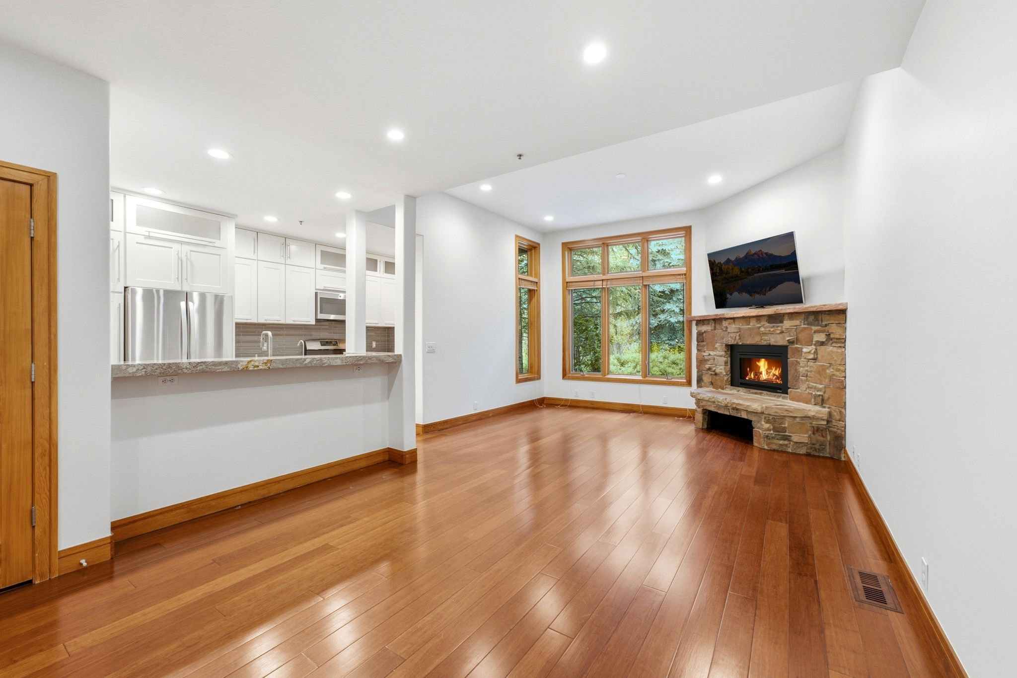 Open-concept living space with warm wood-finish flooring, a stone-clad fireplace, and large picture windows