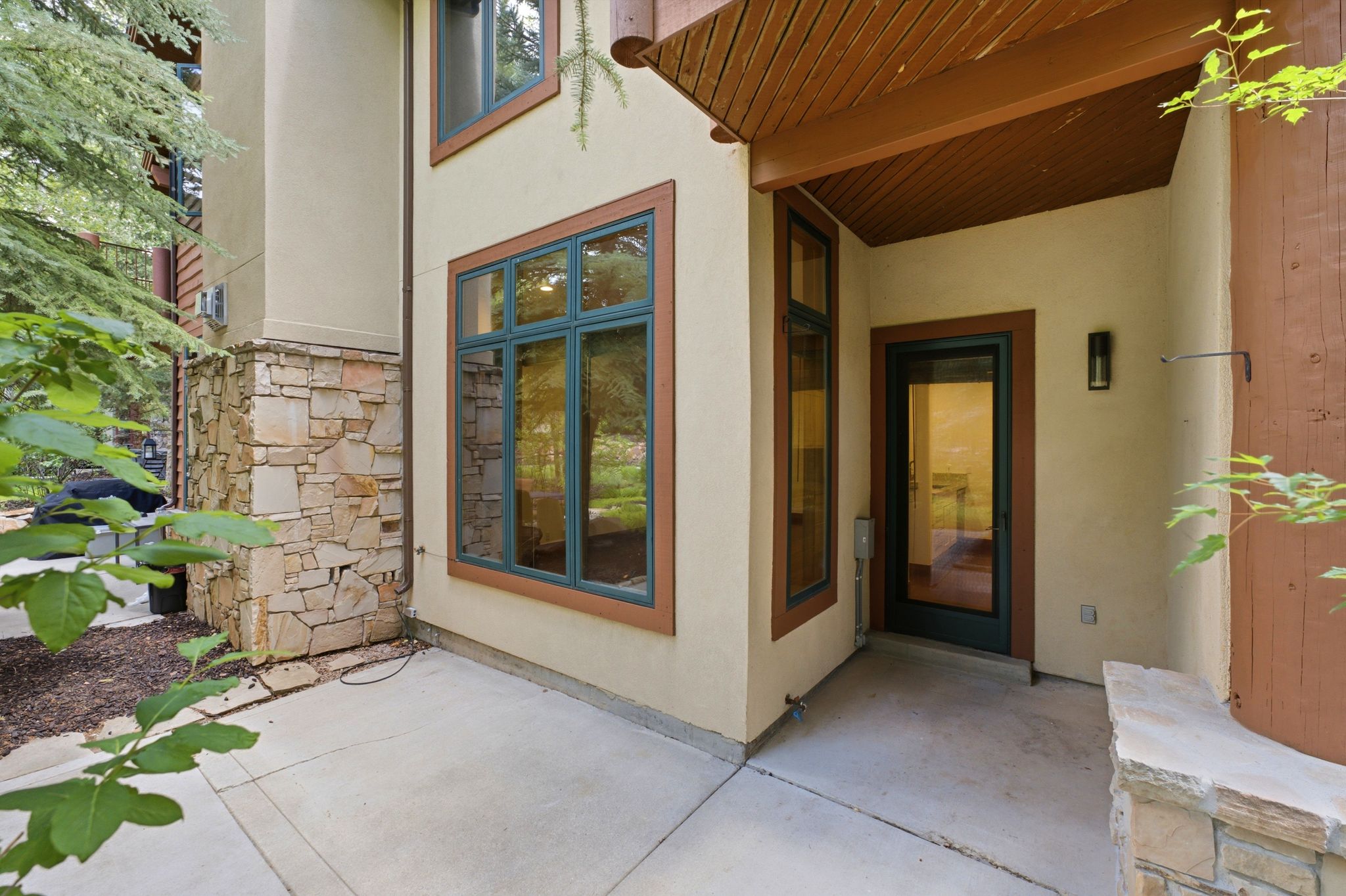 Exterior featuring stucco siding, natural stone accents, large windows with dark frames, a covered entry with wood-finish ceiling, and a concrete patio
