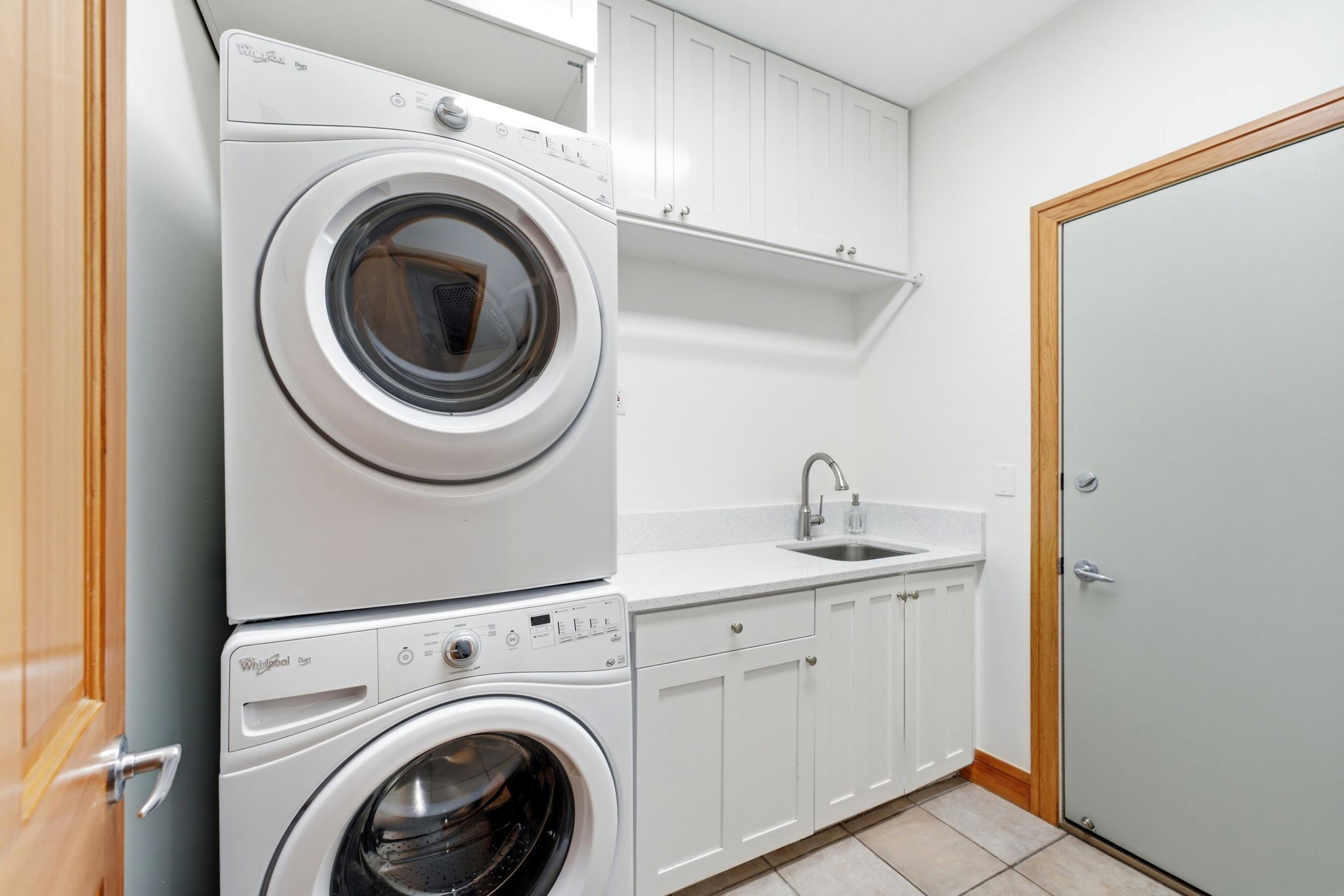 Dedicated laundry area featuring a stacked washer and dryer, white shaker cabinetry, a light-colored countertop with an undermount sink, and durable tile flooring