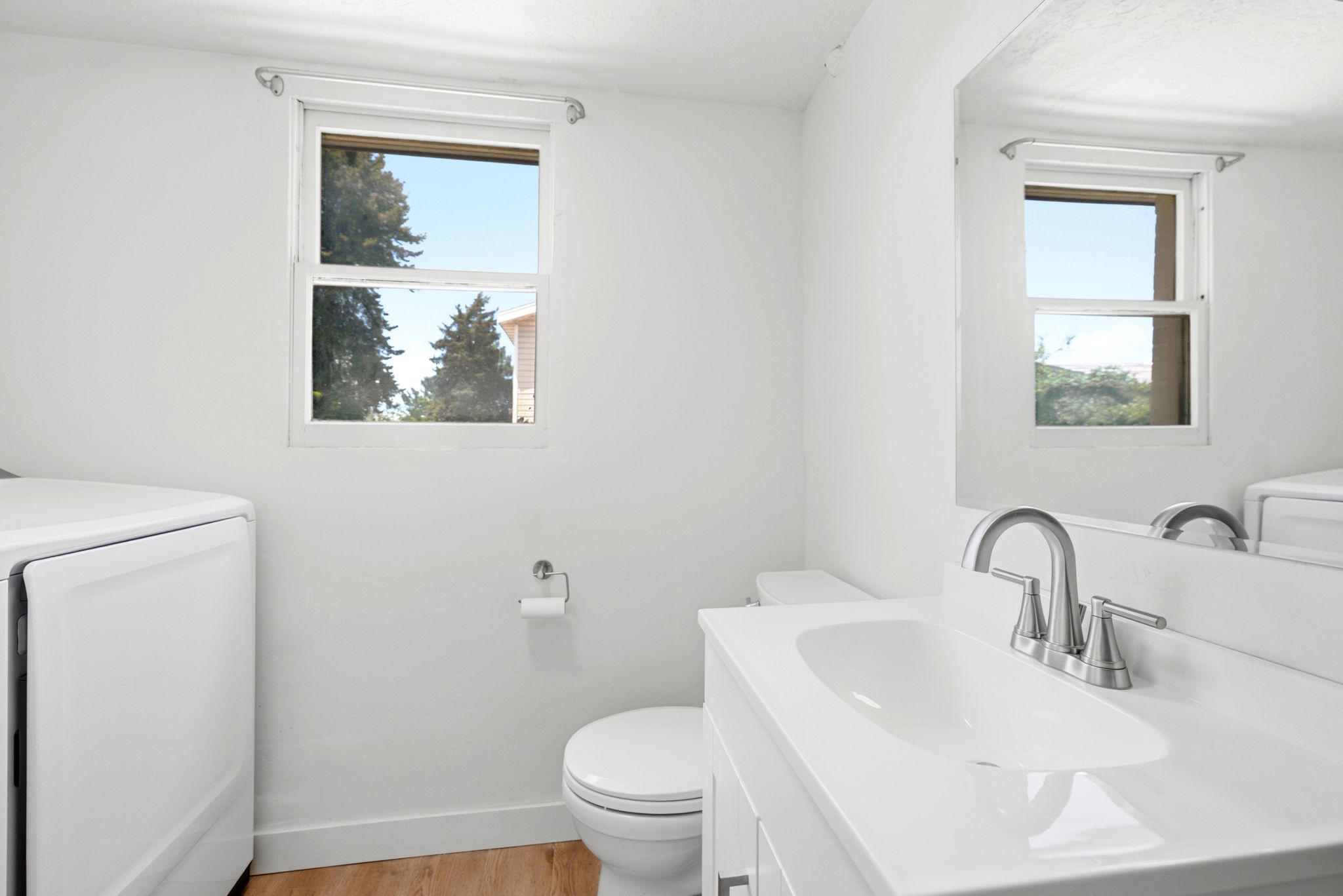 Bathroom featuring a single window, a bright white vanity with an integrated sink and chrome faucet, a wall-mounted mirror, and wood-finish flooring