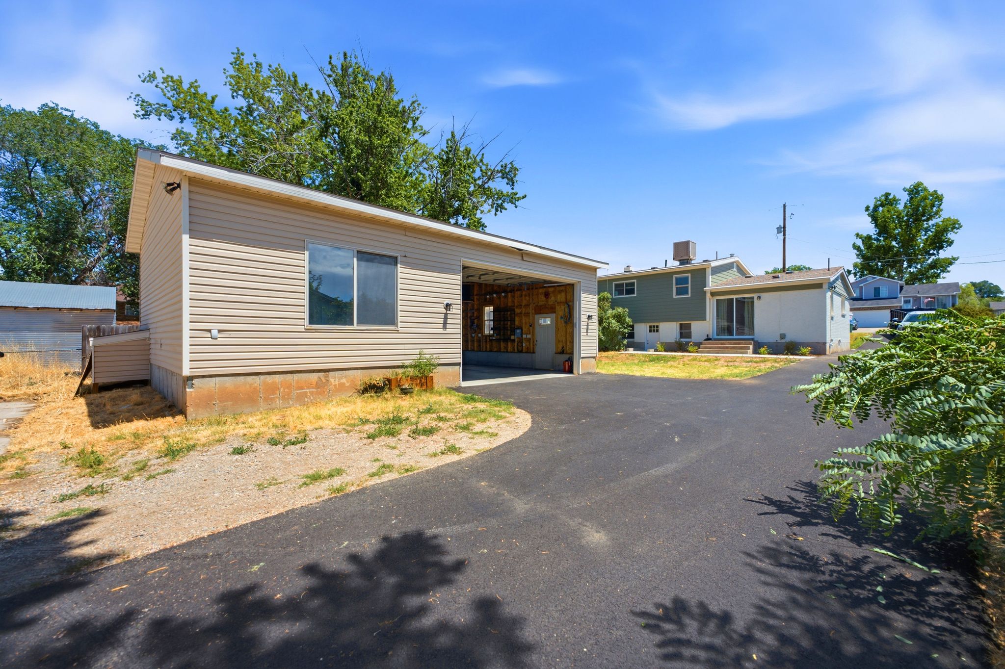 Detached beige vinyl-sided structure with a single window and open bay, featuring a black asphalt driveway