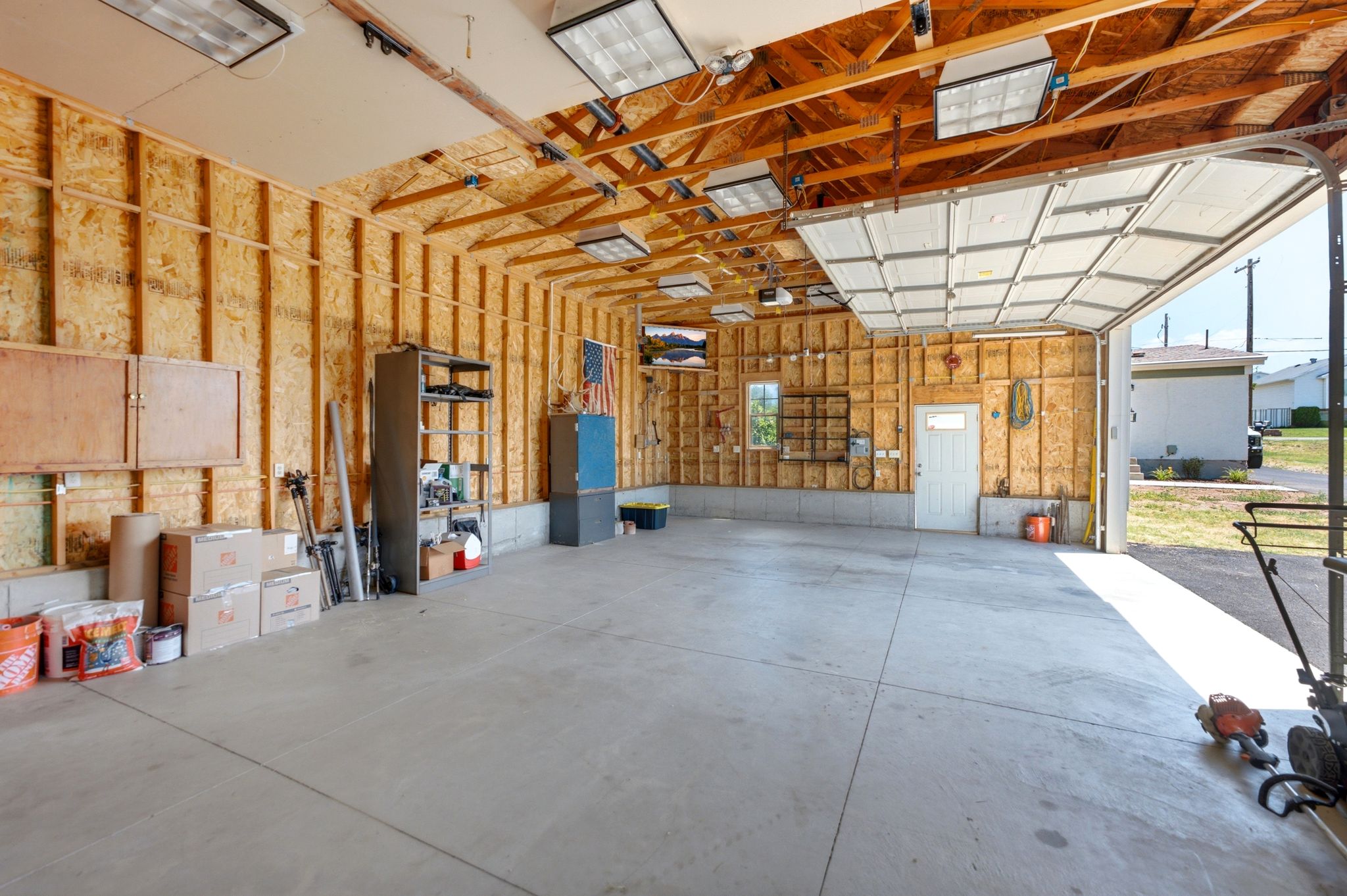 Spacious garage featuring exposed wood framing, multiple fluorescent light fixtures, a white overhead garage door, and a dedicated pedestrian access door