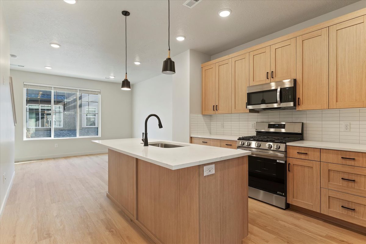 Open concept kitchen featuring an island with a white countertop and undermount sink, pendant lighting, stainless steel appliances, light wood-finish cabinetry, and a horizontal tile backsplash