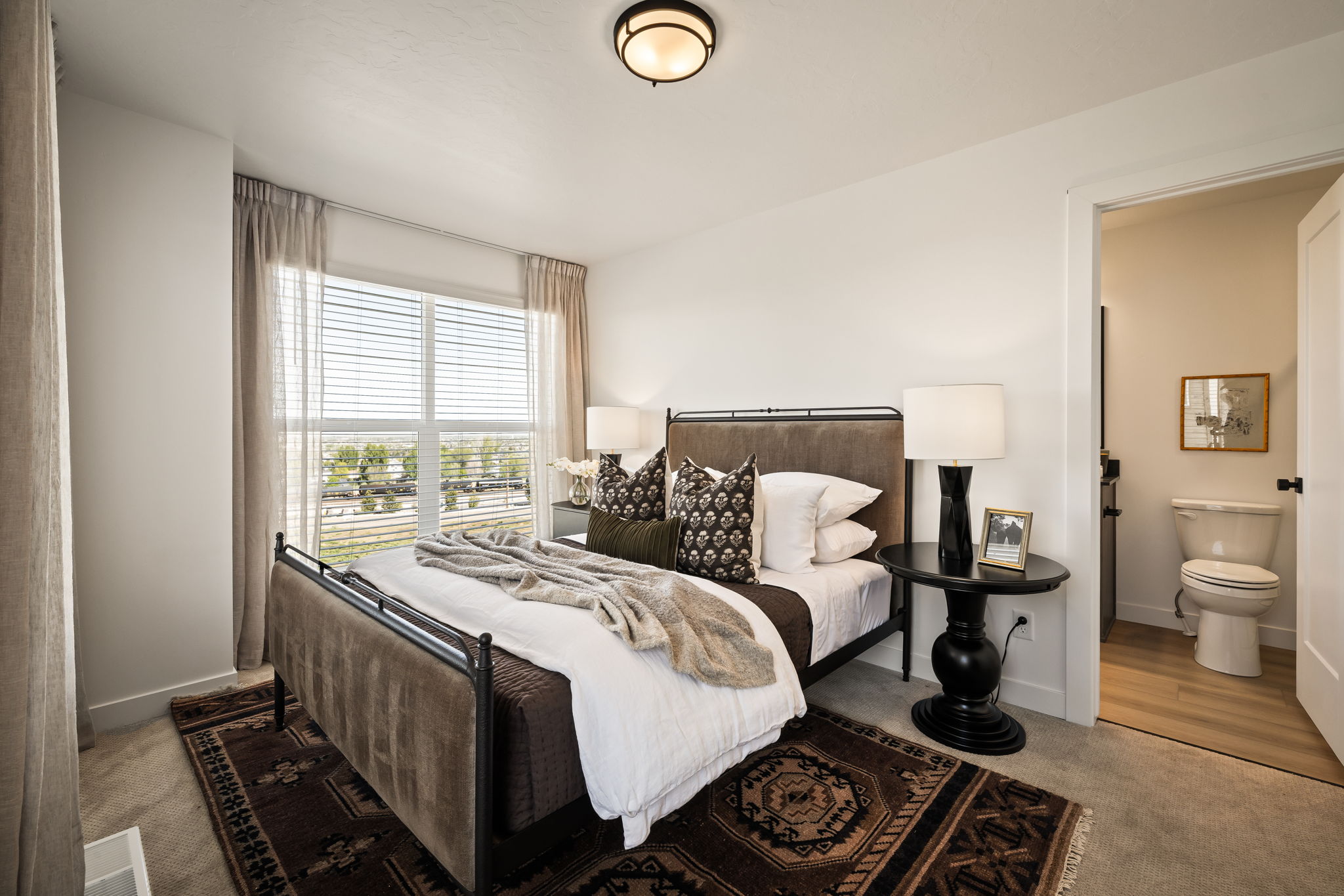 Carpeted bedroom featuring a large window with blinds and drapes, an overhead light fixture, and an entry to a bathroom with wood-finish flooring