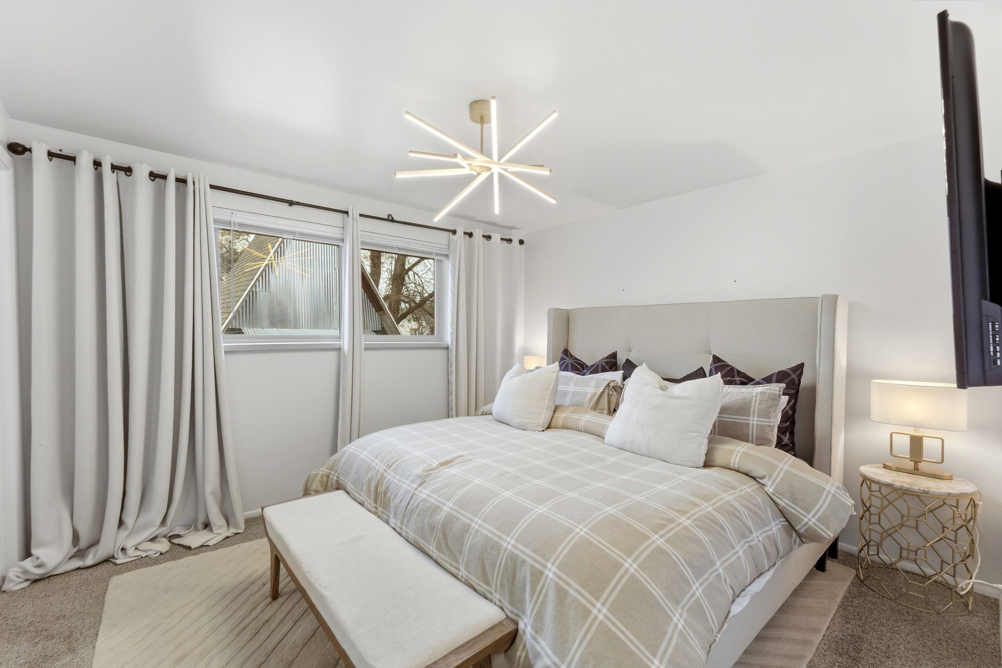 Spacious room featuring two windows with blinds and curtain rods, a modern ceiling light fixture, a built-in wall-mounted television, and a plush upholstered headboard