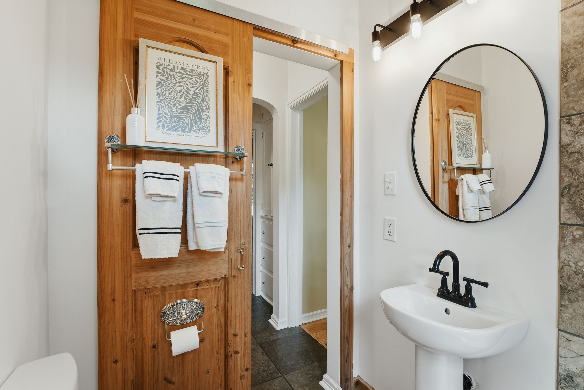 Pedestal sink with black fixture, round mirror, and a wall-mounted light bar