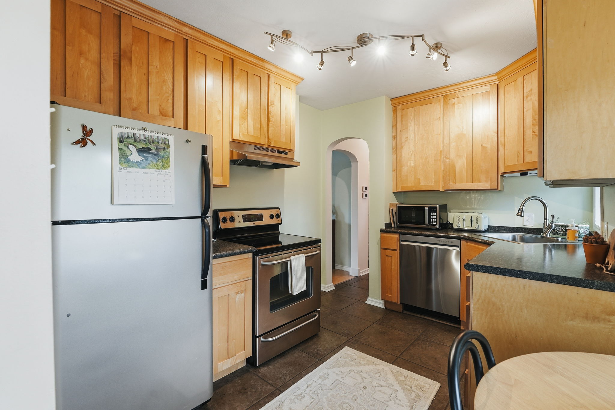 Kitchen featuring natural wood cabinetry, dark countertops, tile flooring, stainless steel appliances, and track lighting