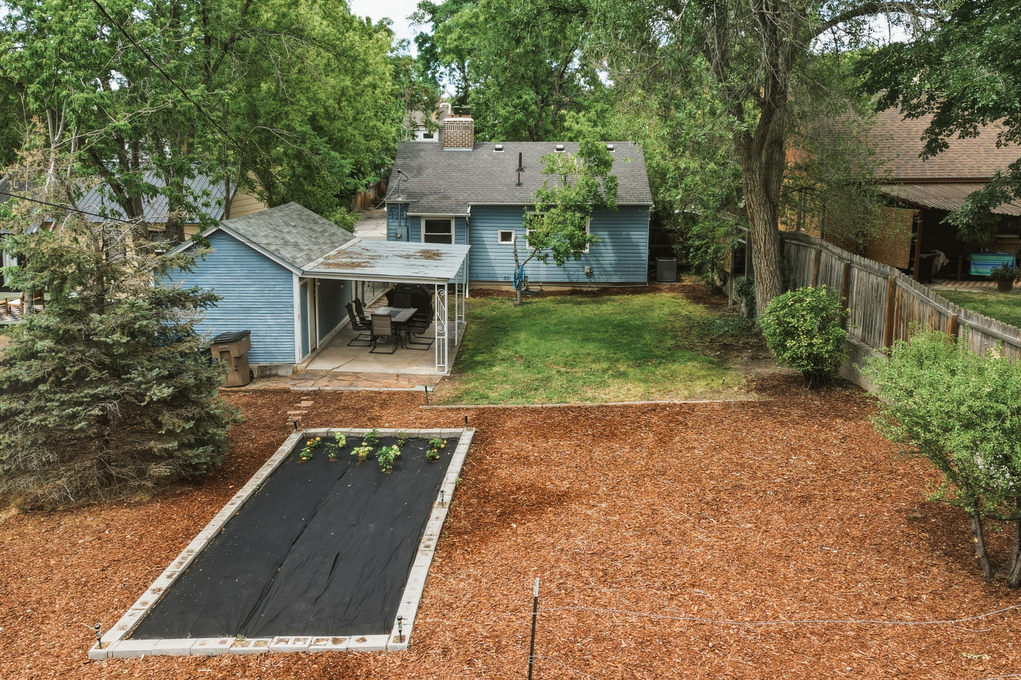Expansive backyard featuring a covered patio, detached shed, raised garden bed with weed barrier, mulched areas, and a grassy lawn
