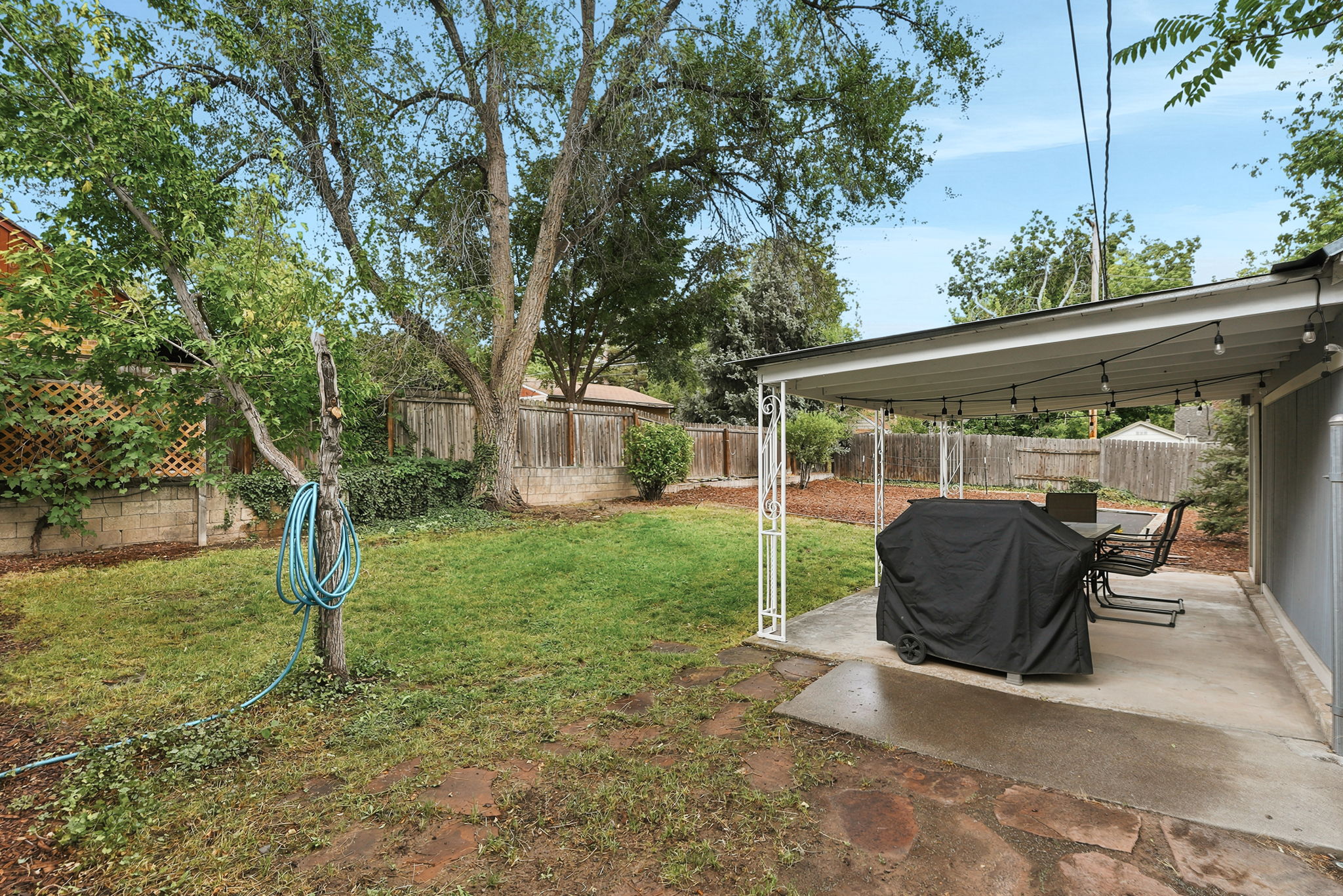 Expansive backyard featuring a covered patio with integrated string lights, a concrete slab, a partial flagstone patio, a wood fence, and mature trees