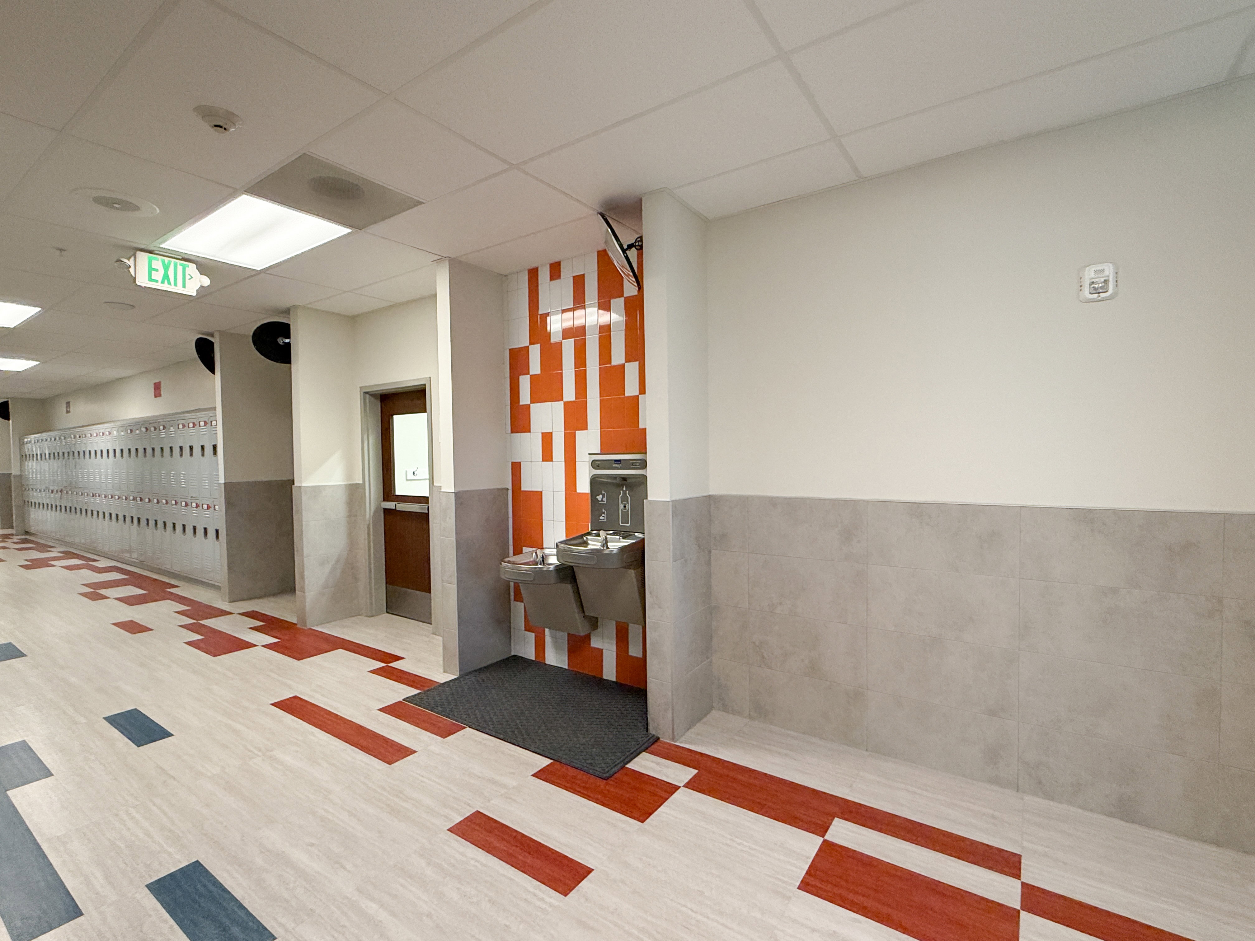 Corridor with light-toned flooring featuring red and blue geometric patterns, complemented by a water fountain station with an orange and white mosaic-tiled accent wall