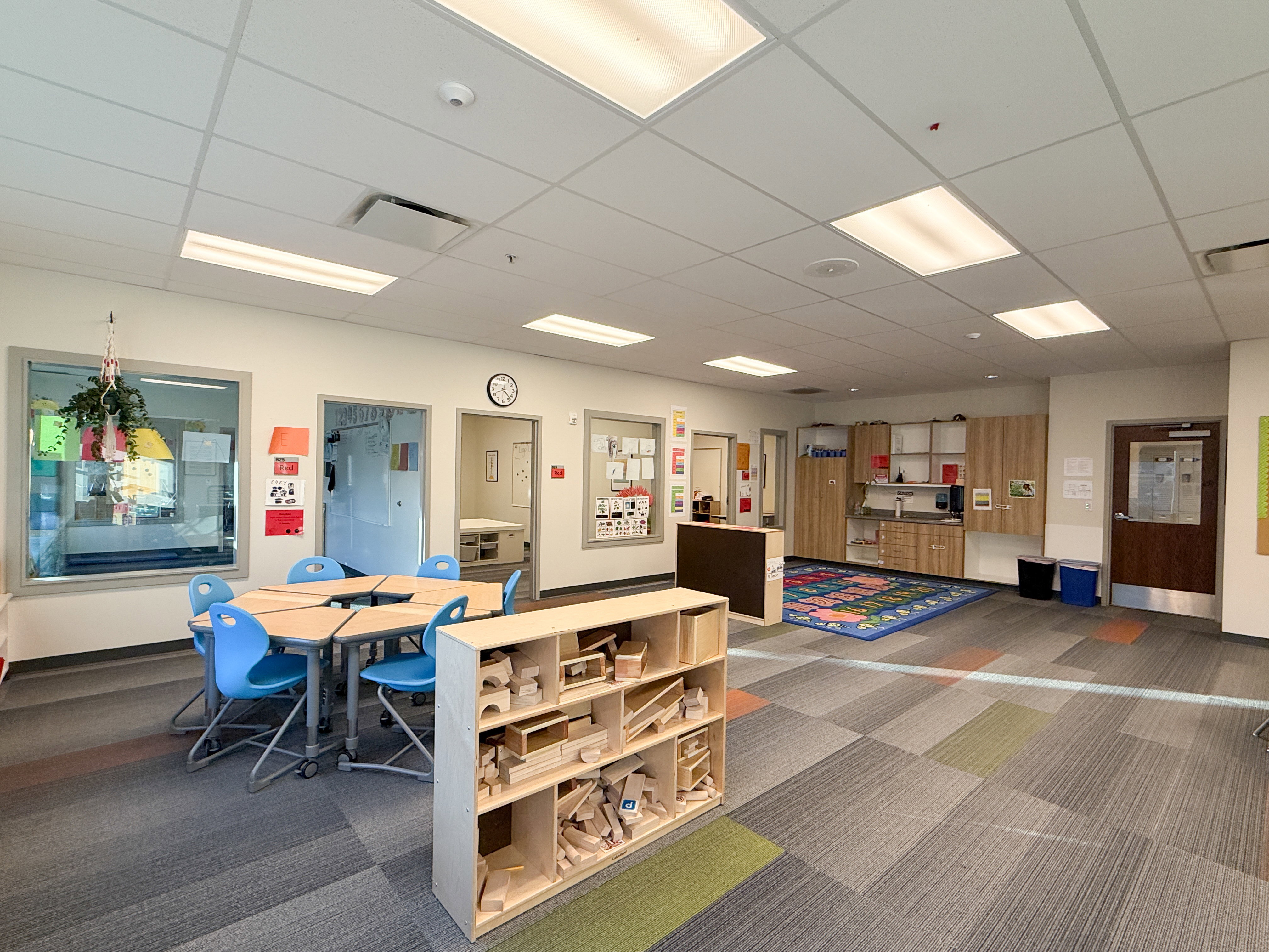 Spacious interior featuring a grid ceiling with integrated lighting, wall-mounted cabinetry, durable modular carpeting, and multiple windows and doorways providing natural light