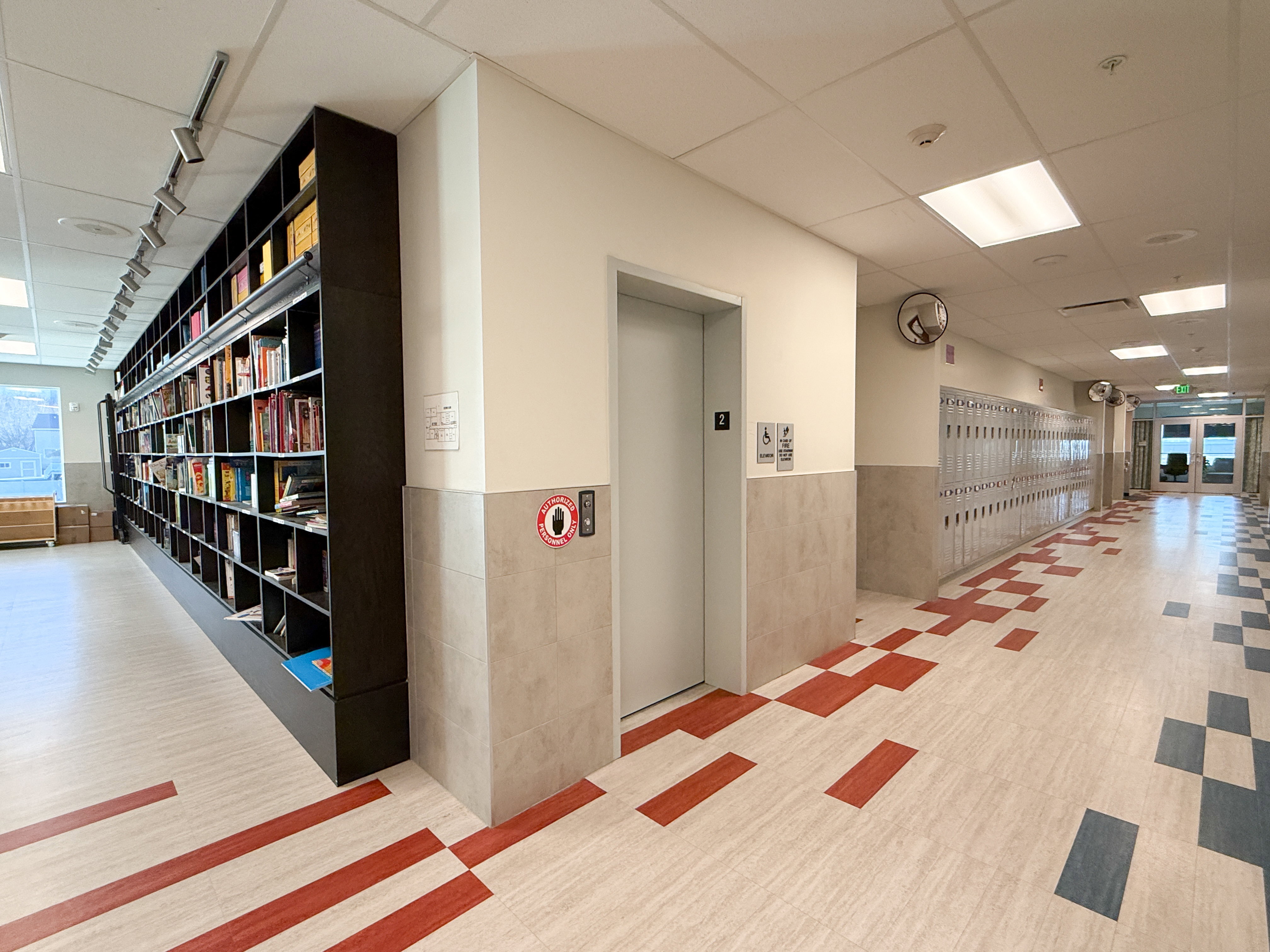 Hallway featuring a built-in dark wood-finish bookshelf with track lighting, a light-colored elevator, and a patterned tile floor